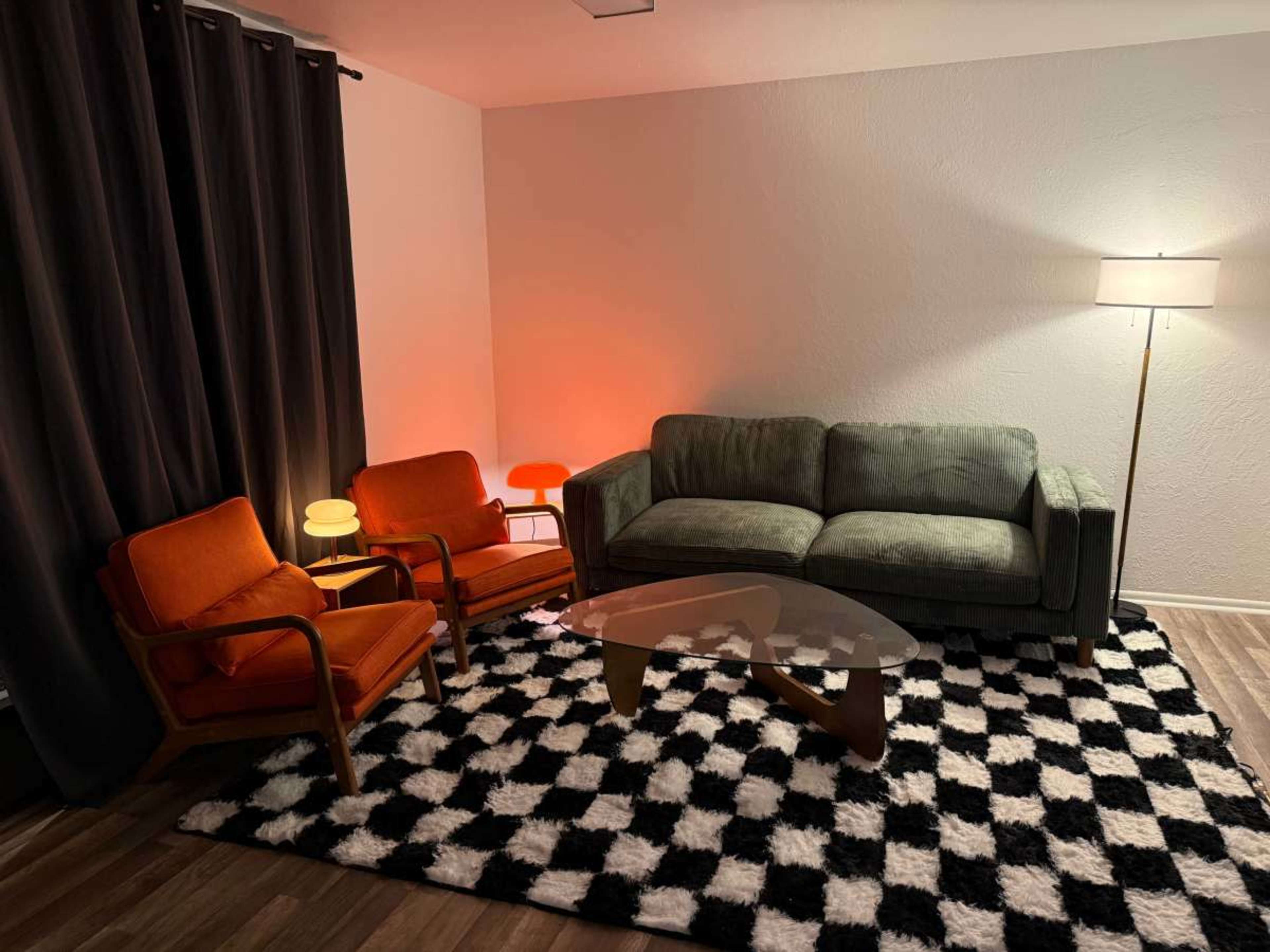 The image shows a living room with a green sofa, two orange chairs, a coffee table, and a black and white checkered rug.