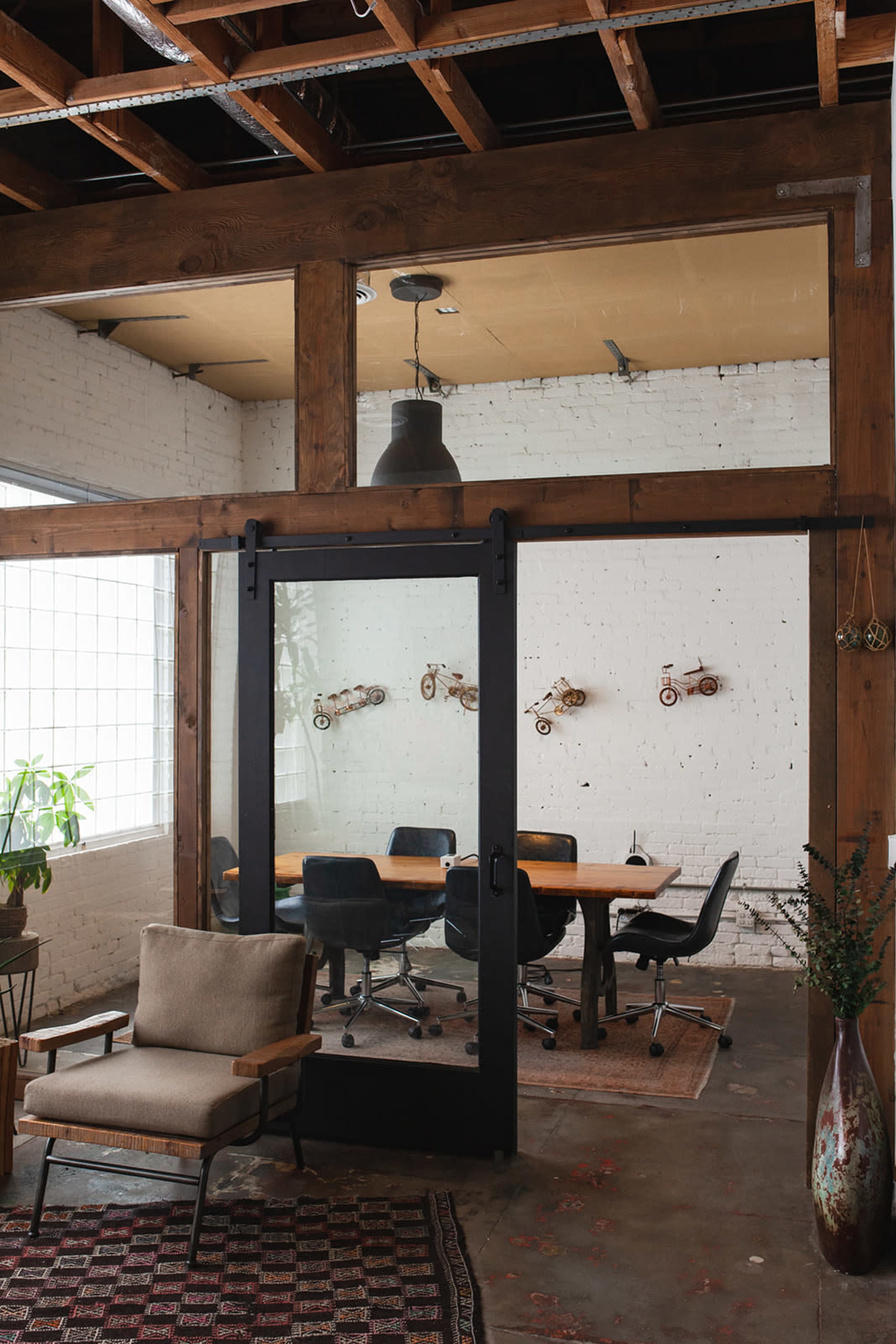 A modern office space featuring a wooden framed glass door that leads to a conference room with a large table and black chairs, surrounded by exposed brick walls.
