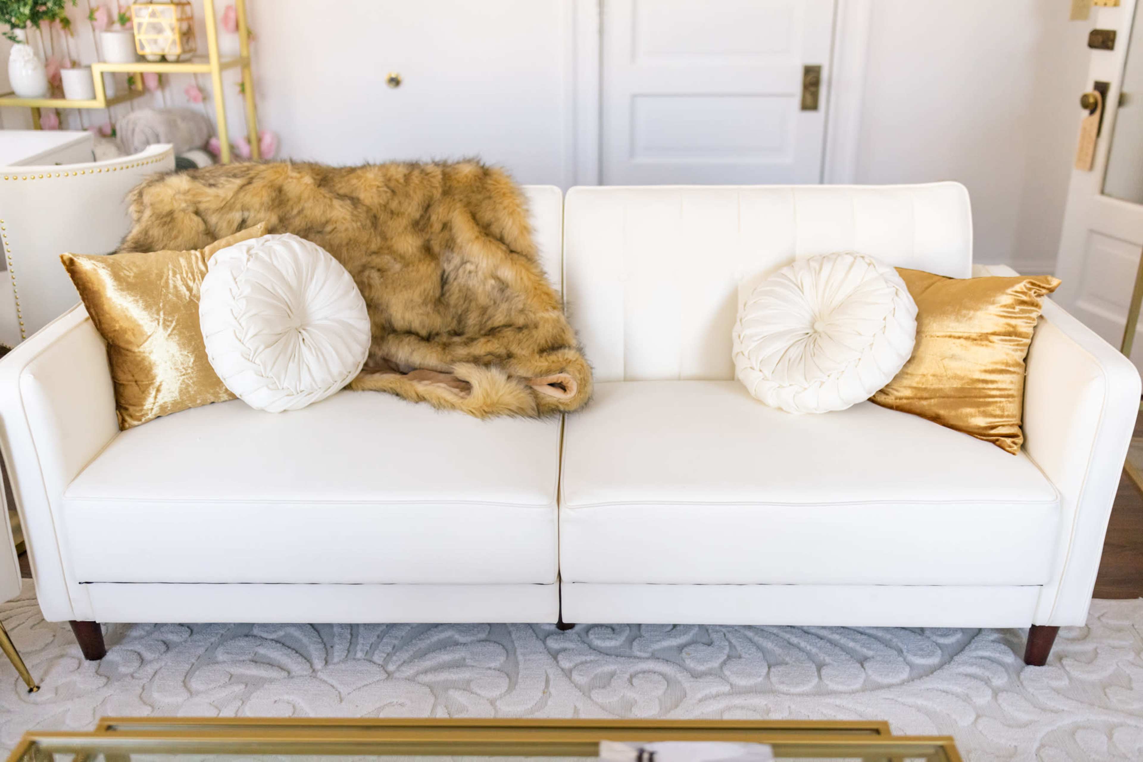A white sofa with decorative pillows and a fur throw is positioned in a bright room with a light-colored rug.