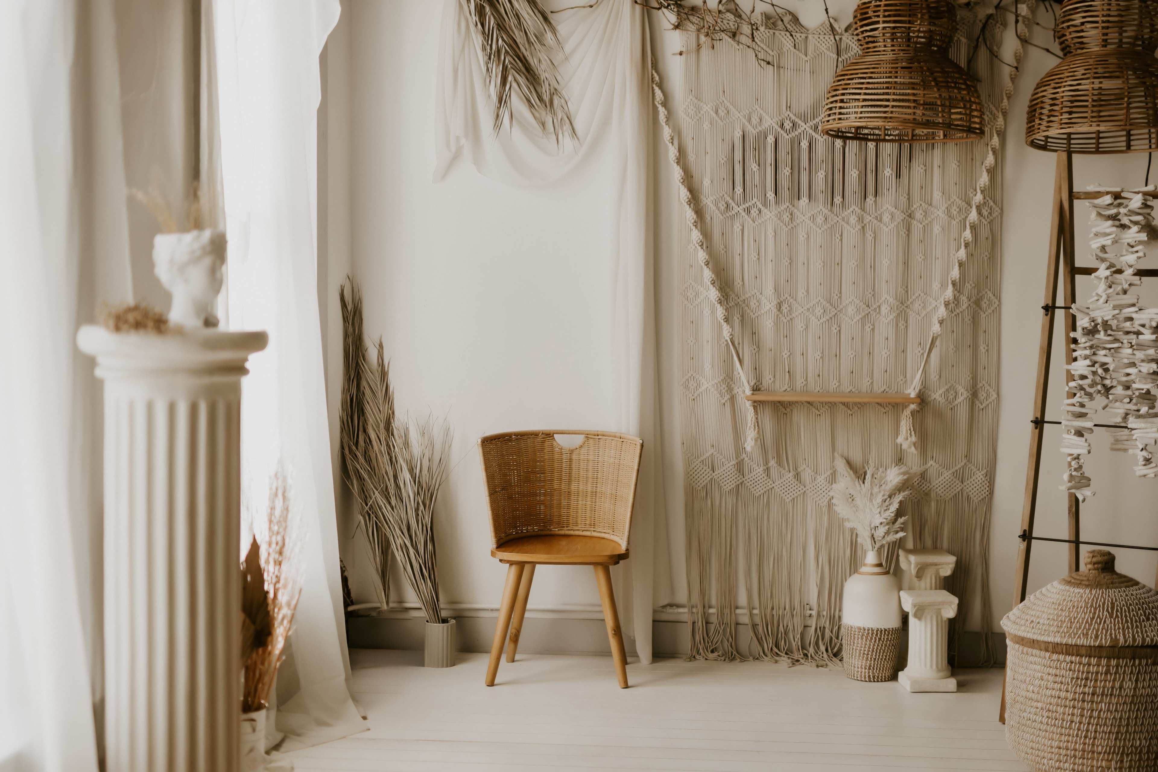 The image shows a minimalist interior space featuring a woven chair, decorative wall hangings, and various plants arranged aesthetically.