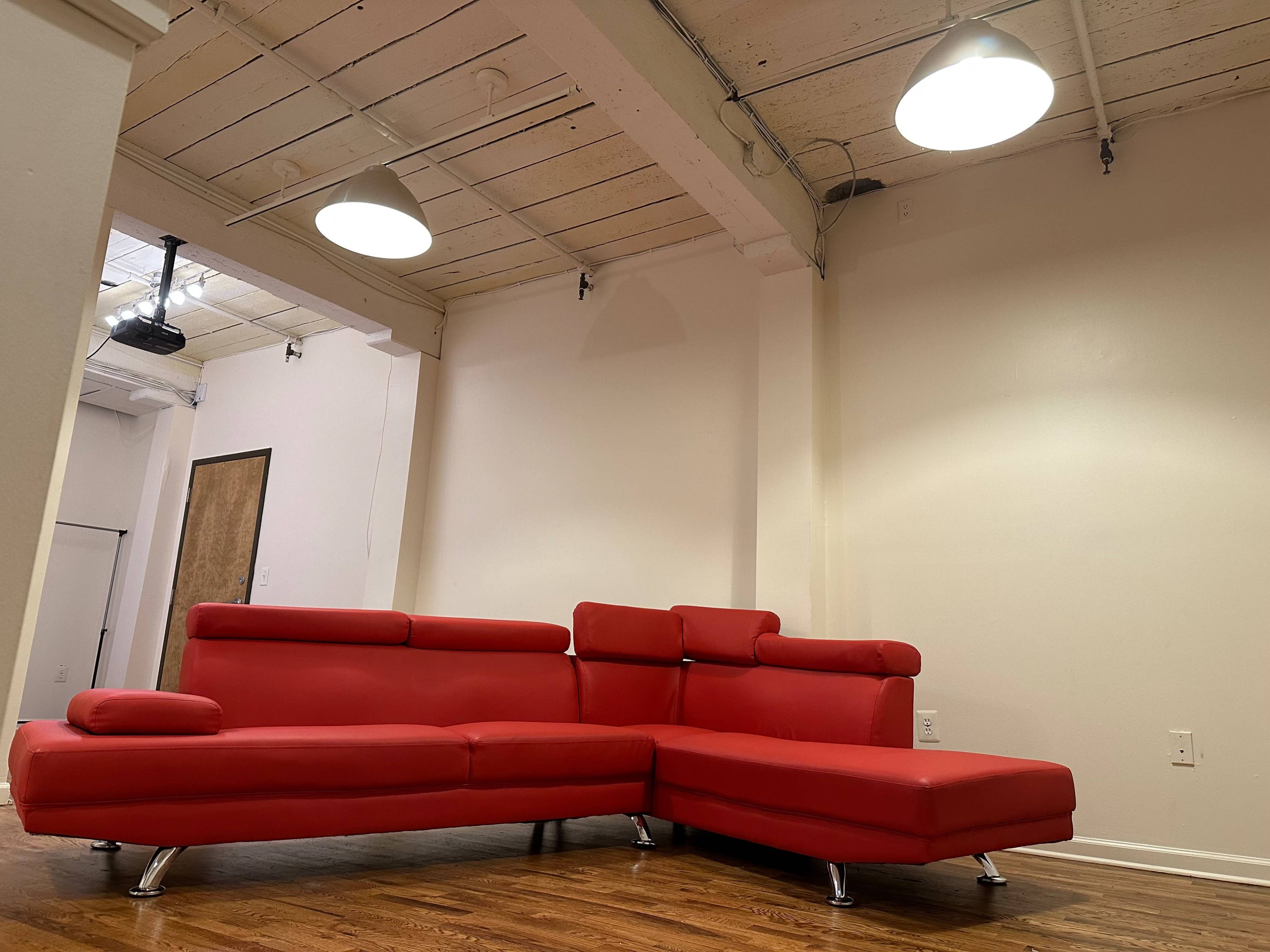 The image shows a red sectional sofa placed in a spacious, minimally decorated room with wooden flooring and exposed ceiling beams.