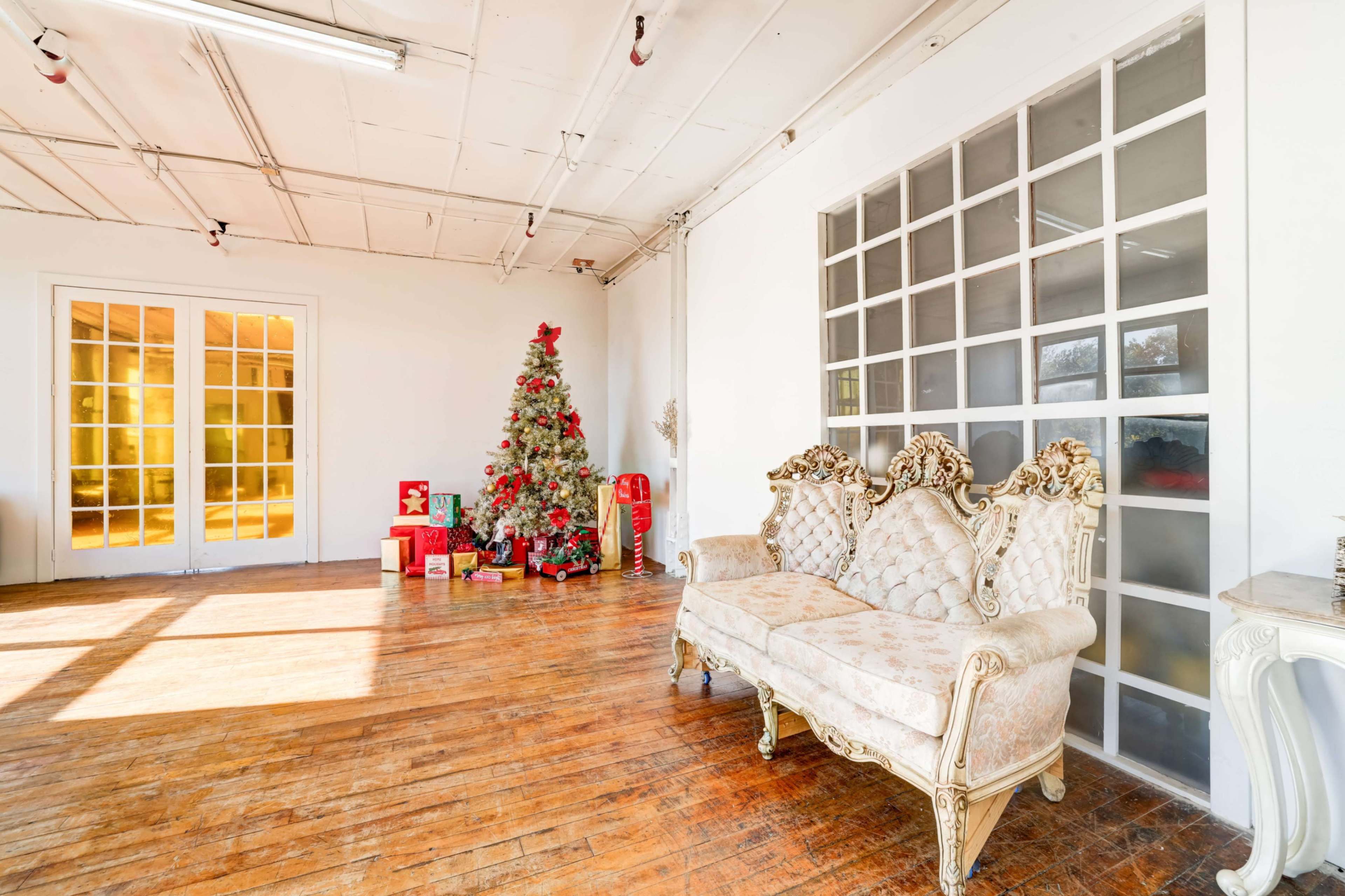A decorated Christmas tree stands next to a vintage couch in a bright, open room with wooden flooring and large windows.