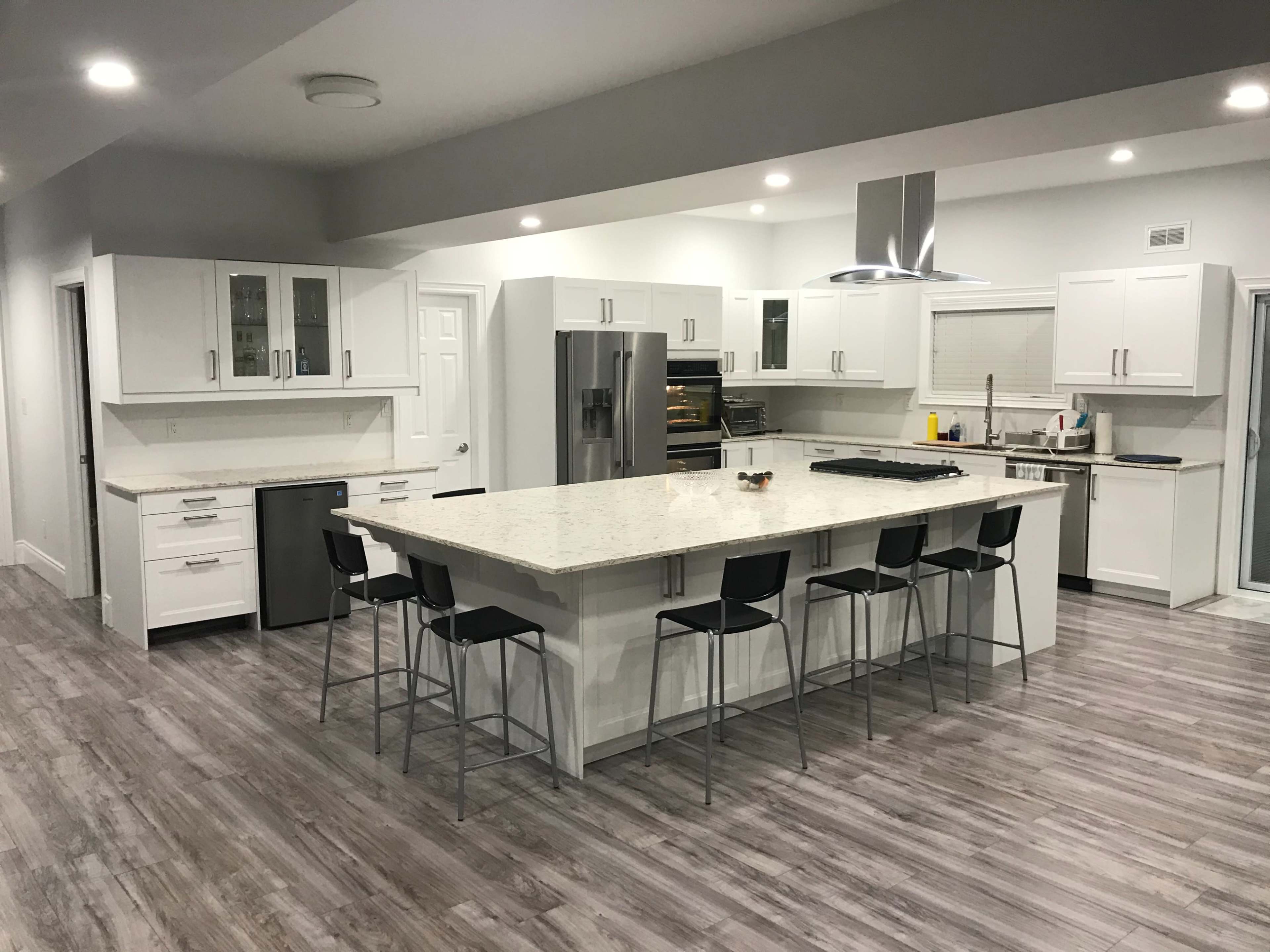 A modern kitchen with a large central island, white cabinetry, stainless steel appliances, and gray wood-like flooring.