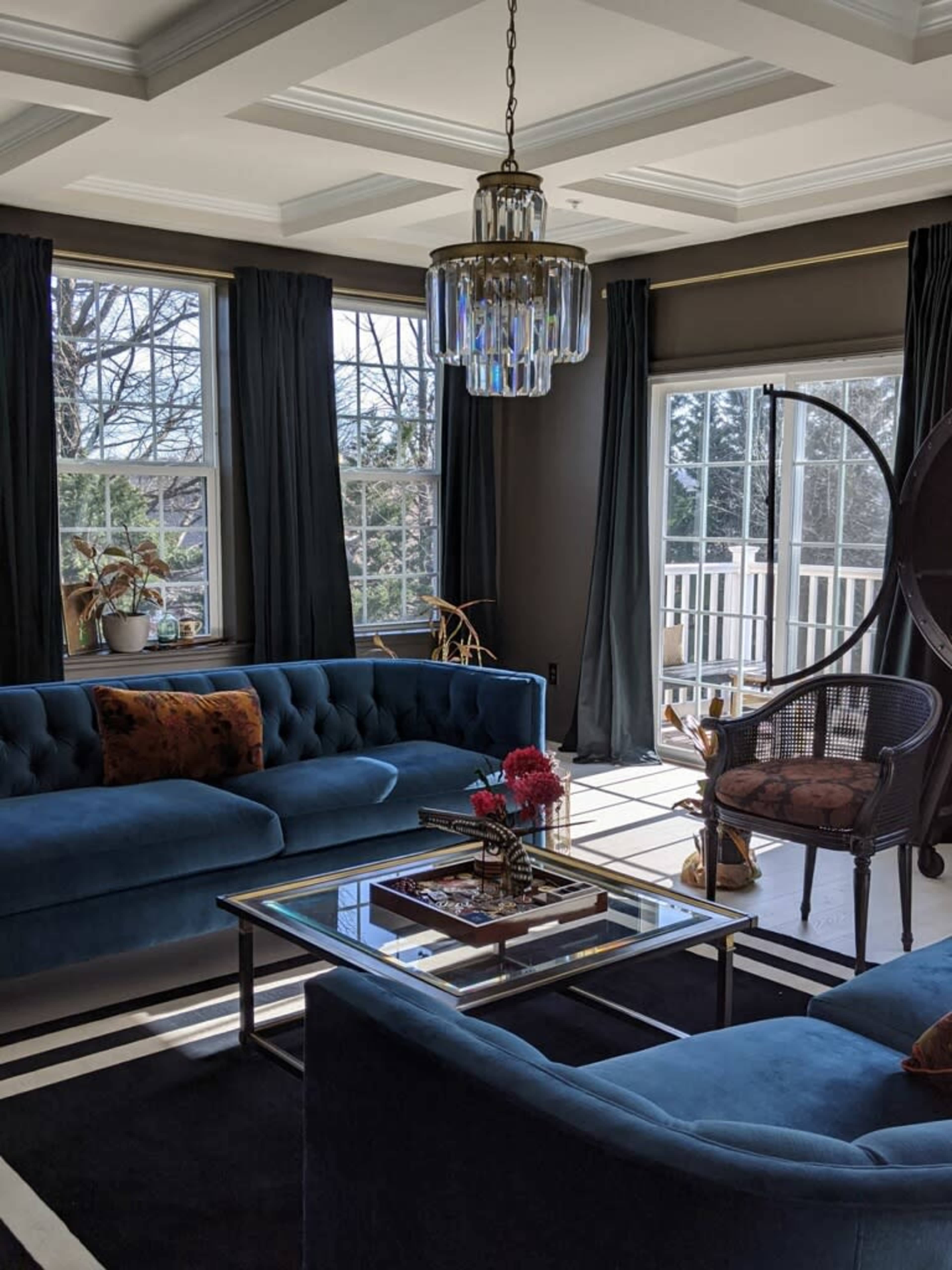 A bright living room features blue sofas, a glass coffee table, and a modern chandelier, with large windows letting in ample natural light.