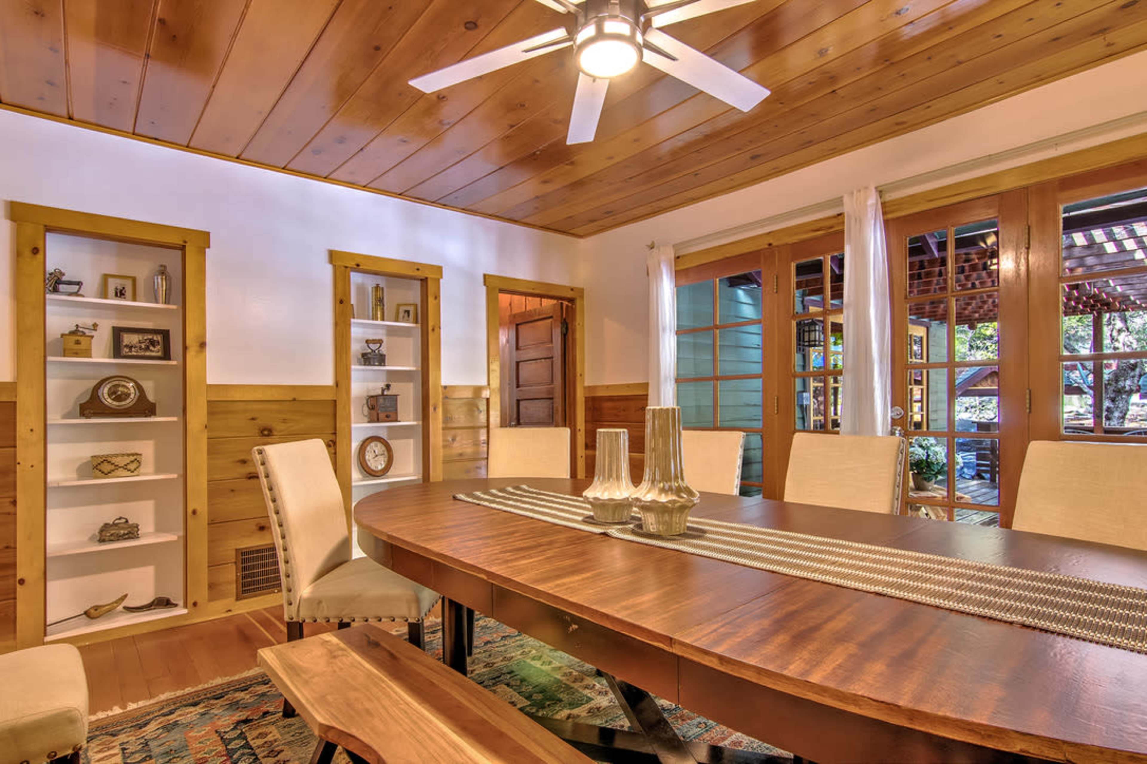 A wooden dining room features a large oval table surrounded by upholstered chairs, with shelves displaying decorative items and large windows opening to an outdoor view.