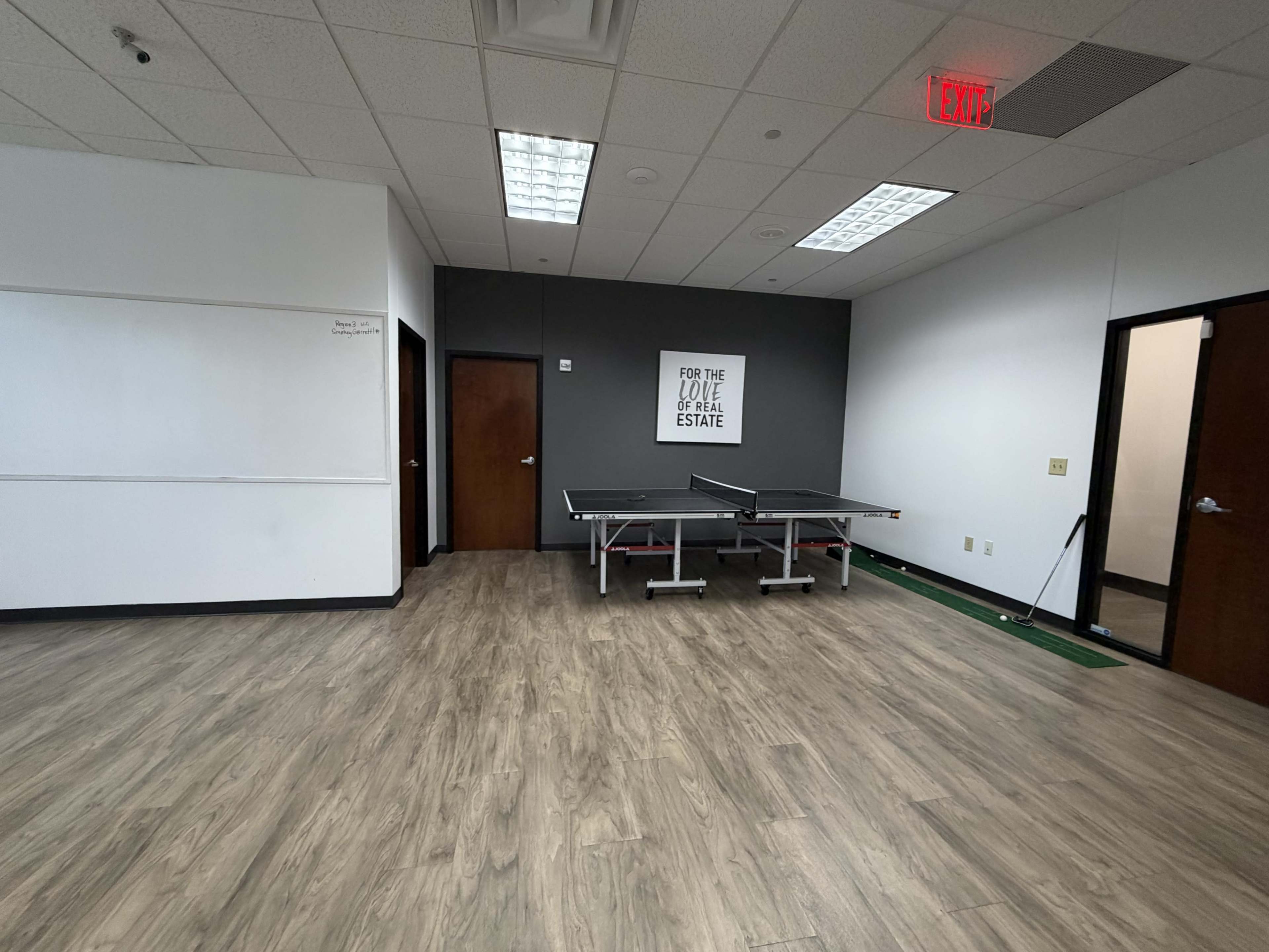 The image shows a spacious room with a table tennis table in the center, whiteboard on the left, and two doors on the right side.