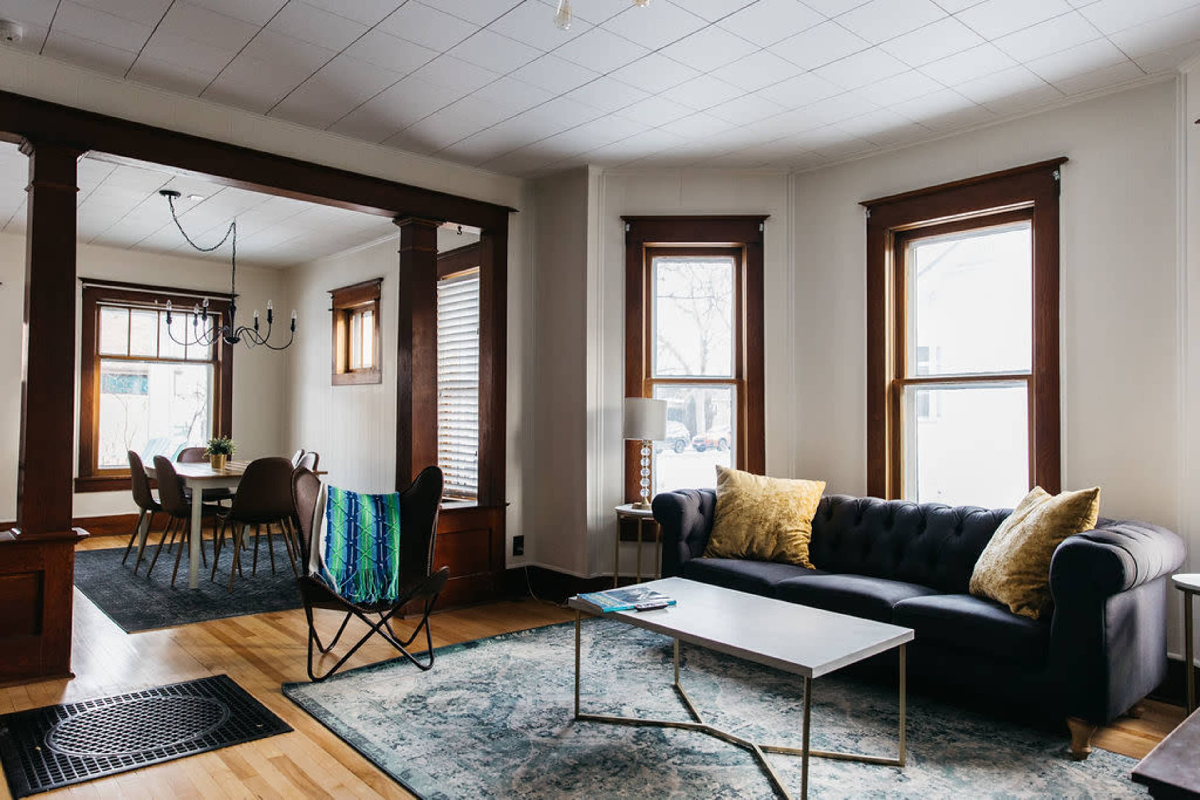 A living room with a dark sofa, a coffee table, and a chair, adjacent to a dining area with a table and chairs, illuminated by natural light through large windows.