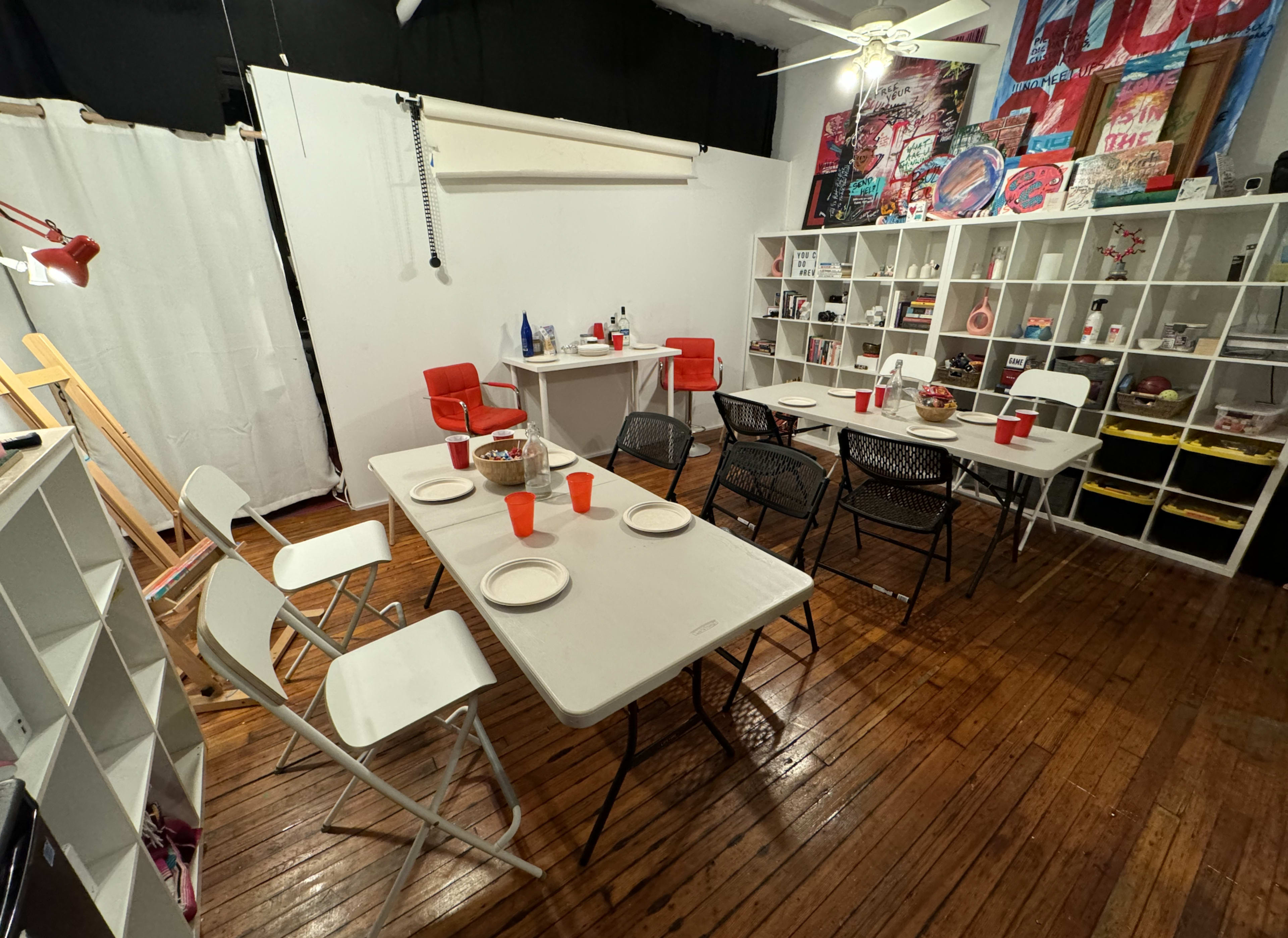 The image shows a room with several folding tables and chairs arranged for a dining setup, along with storage shelves containing various items.