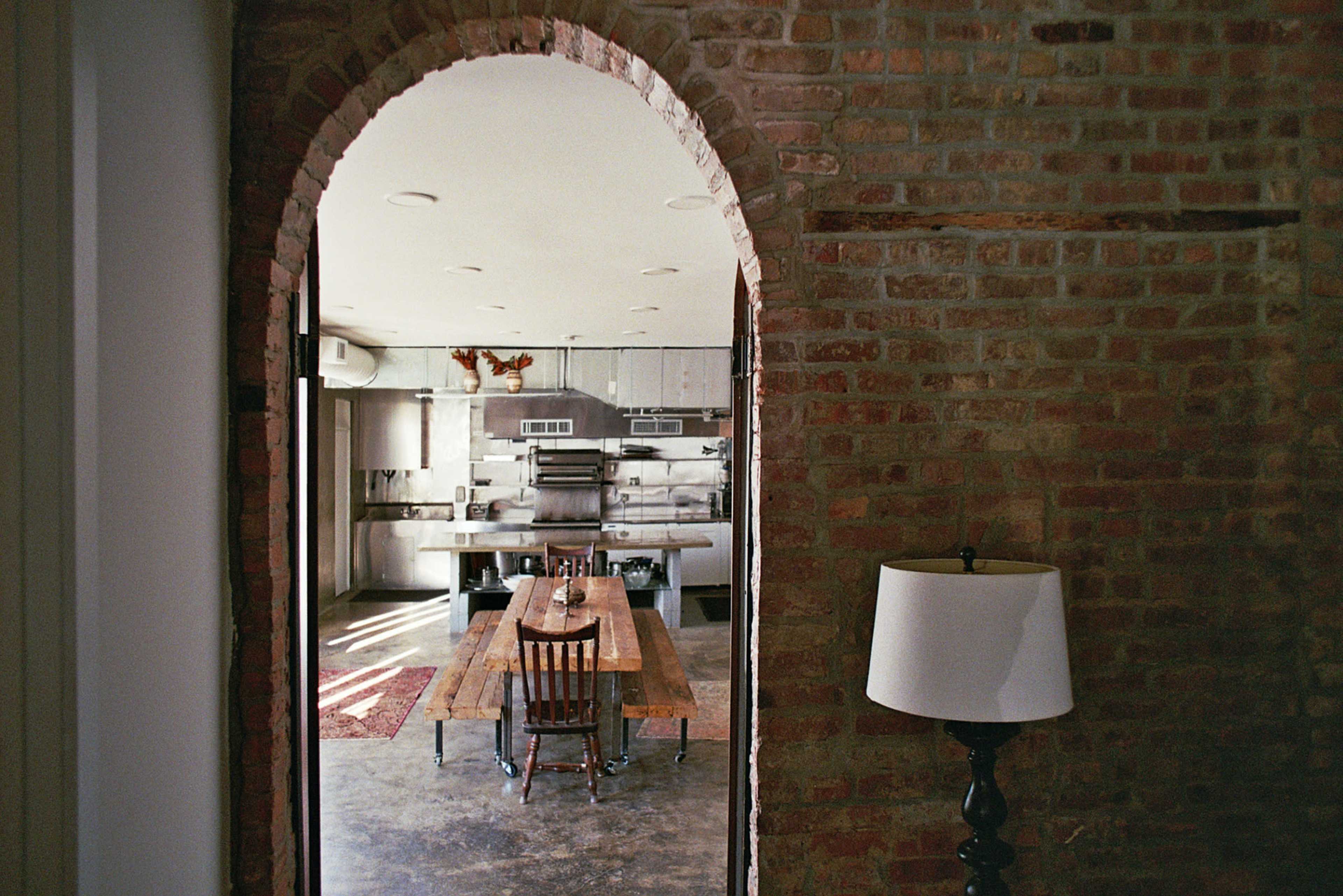 The image shows a brick-walled room with an arched doorway leading to a kitchen area featuring a long wooden table and chairs.