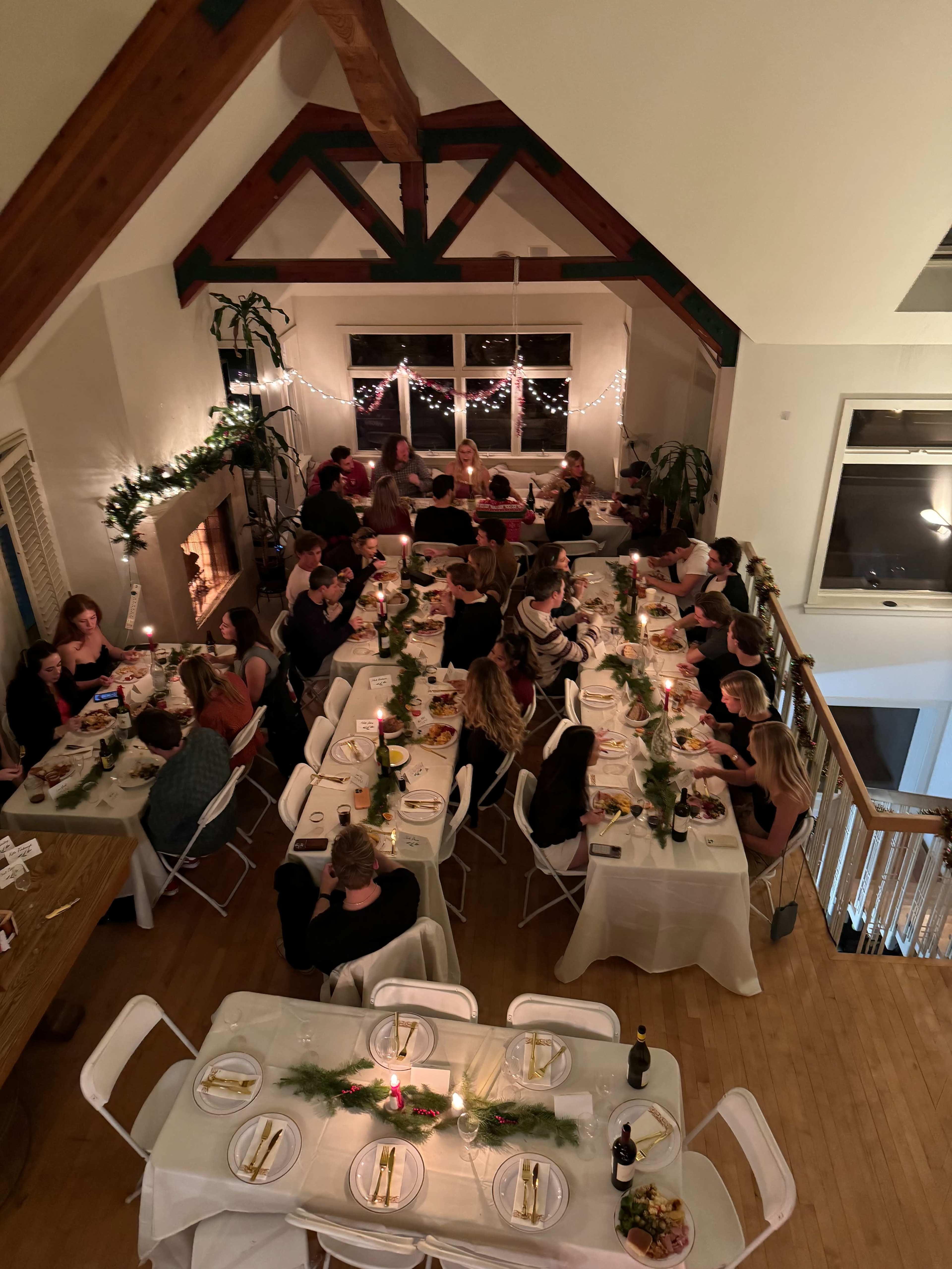 A large group of people is gathered around several long tables set for a dinner in a warmly lit room with wooden beams and decorative greenery.