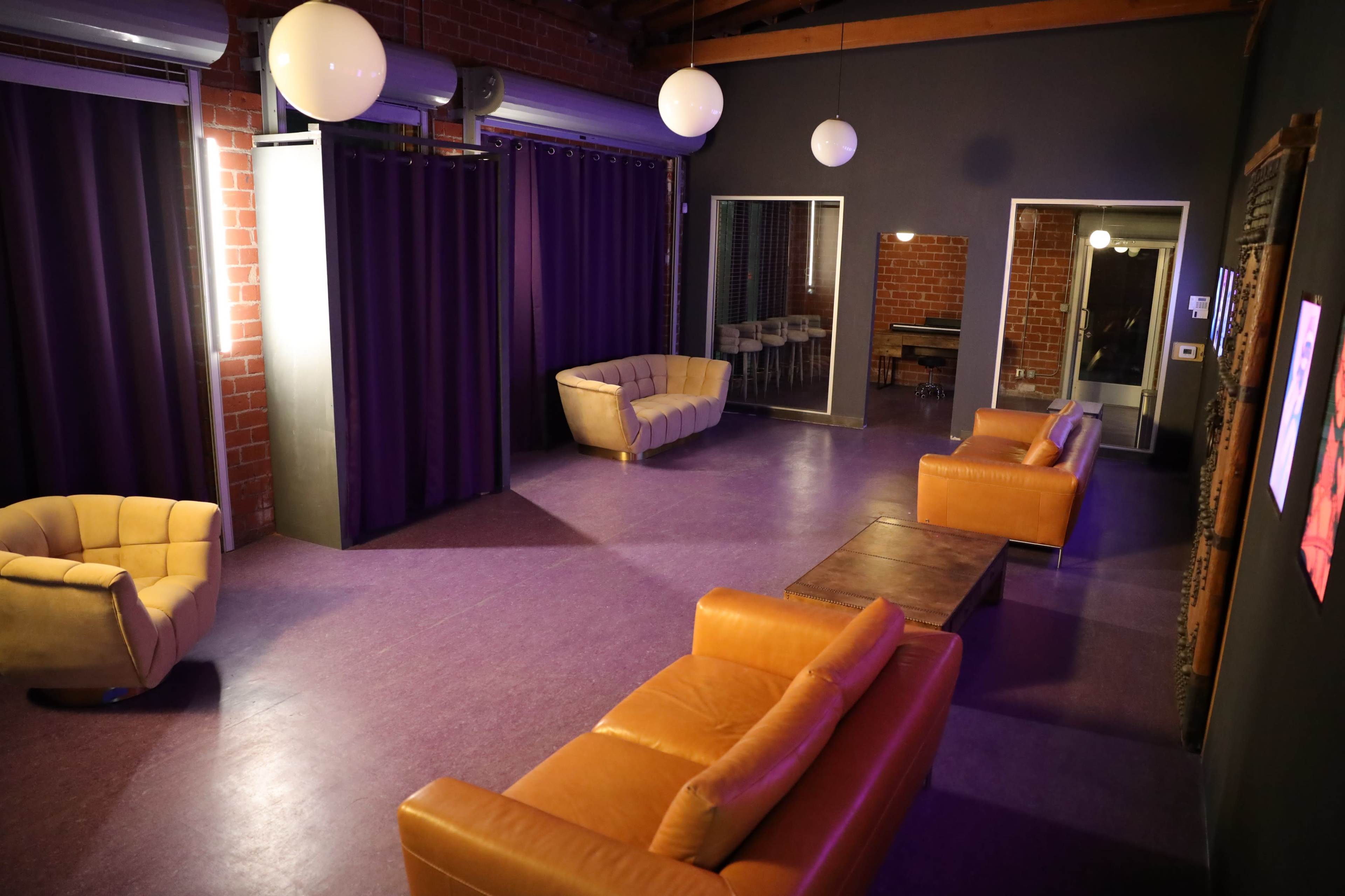 The image shows a spacious lounge area with orange sofas, purple curtains, and wooden furniture under round hanging lights.