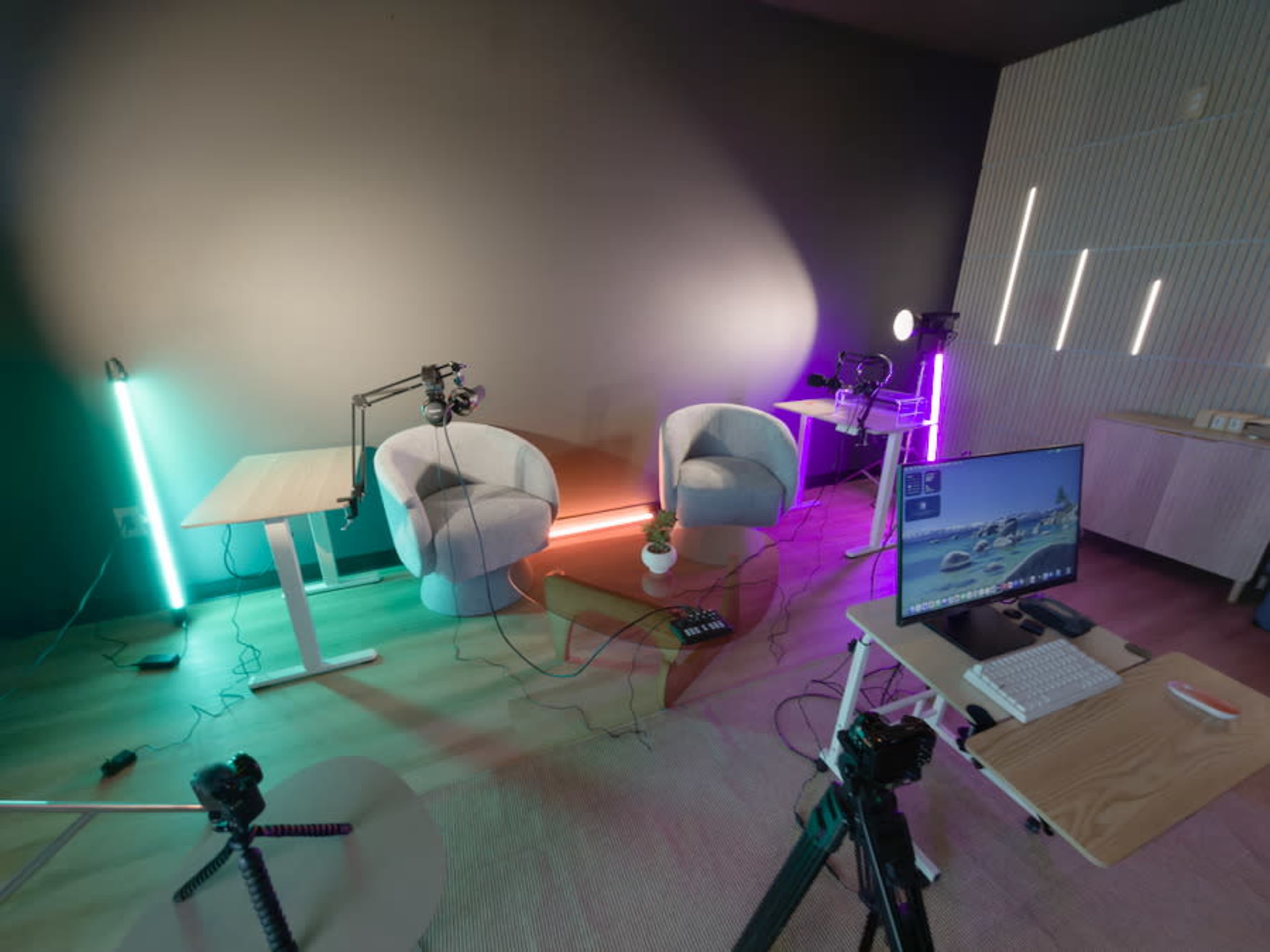 The image shows a modern studio setup with two chairs, colorful lighting, a computer monitor on a desk, and camera equipment positioned on tripods.