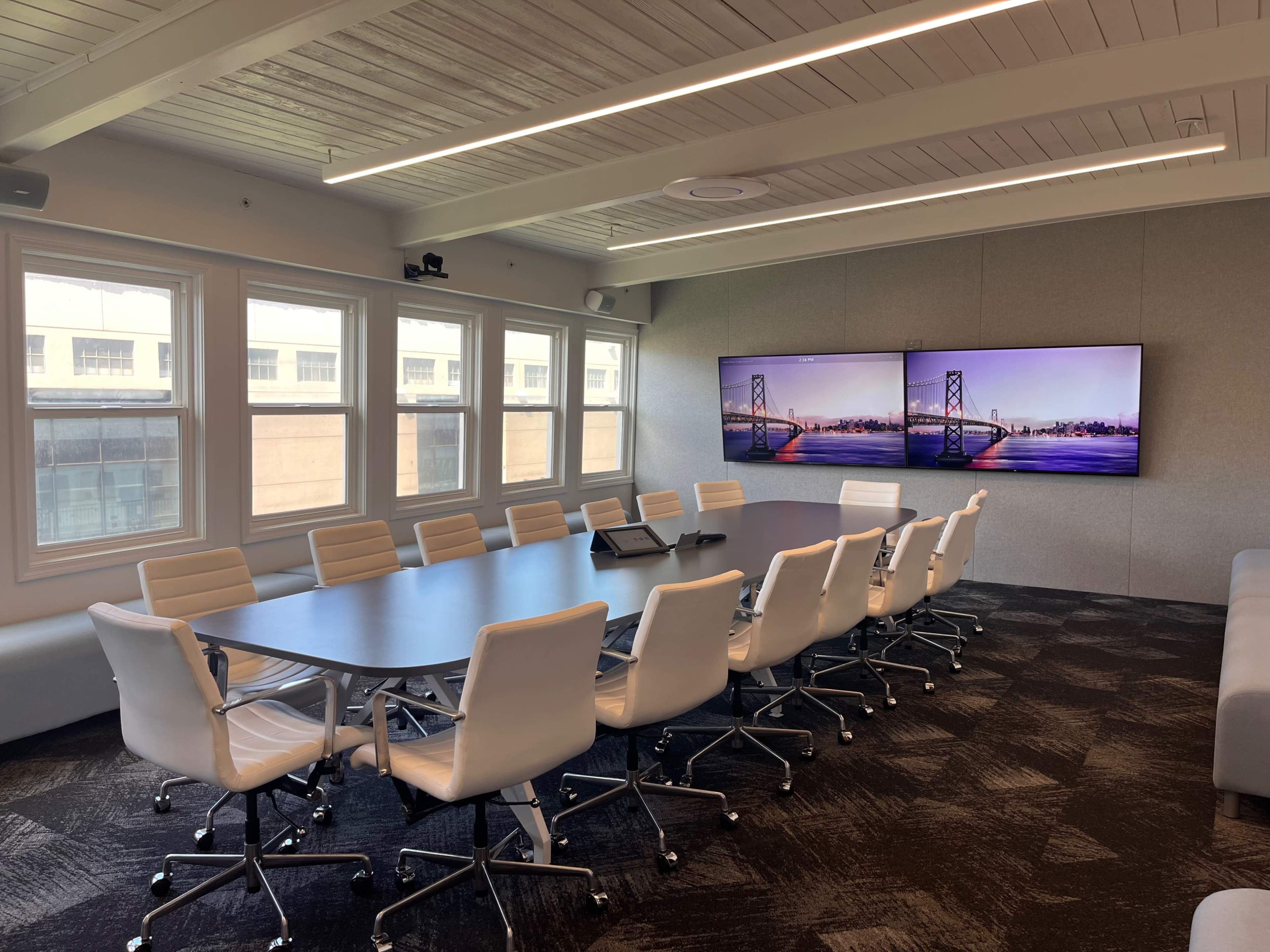 A modern conference room features a large table surrounded by white chairs and two large screens displaying an image of a bridge.