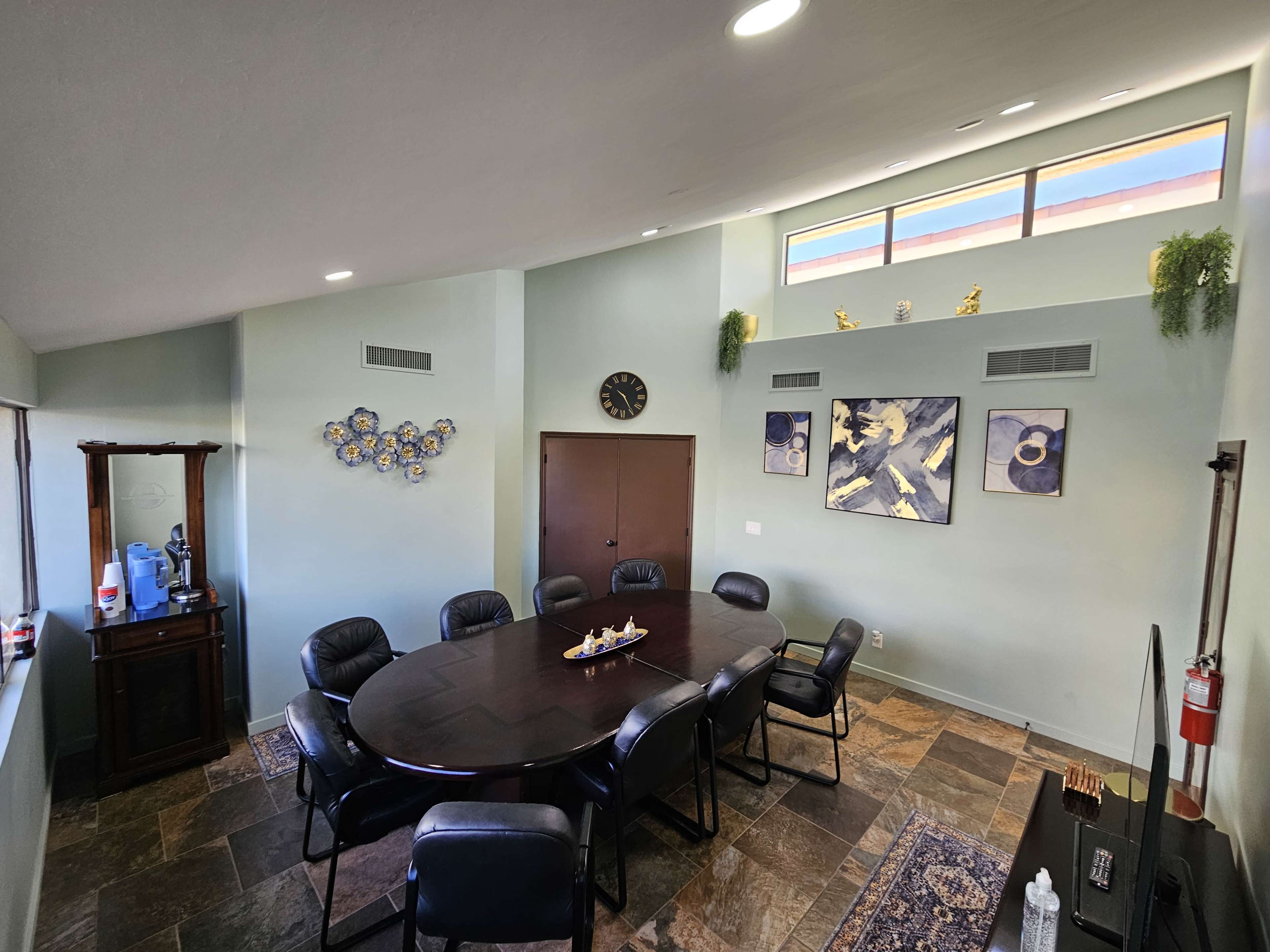 The image shows a modern conference room with a large round table surrounded by black chairs, decorated with wall art and a clock.