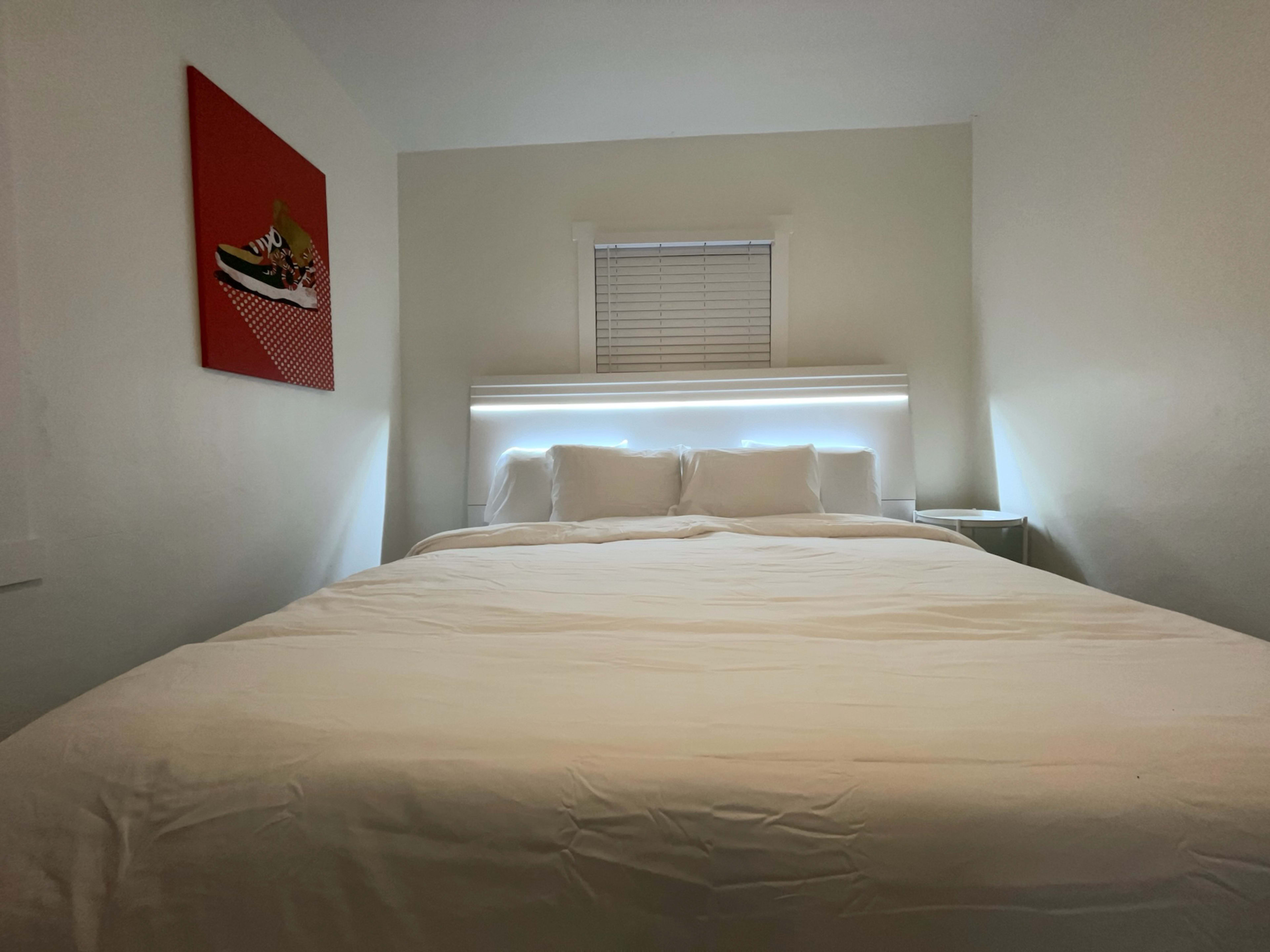 A neatly made bed with white bedding in a well-lit room featuring a red artwork on the wall.