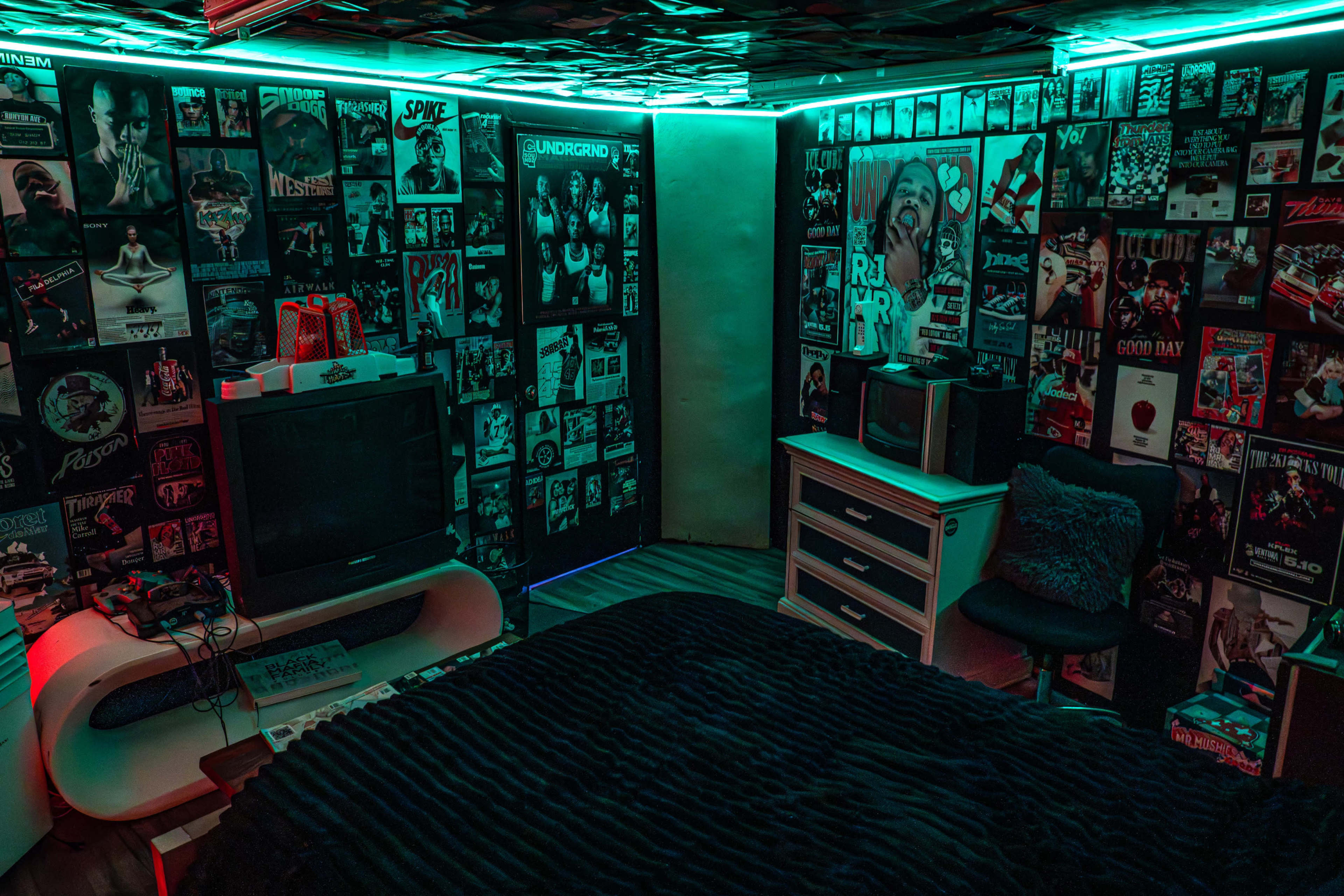 The room features a bed with a dark blanket, two televisions, and walls covered in posters, illuminated by neon blue and green lights.