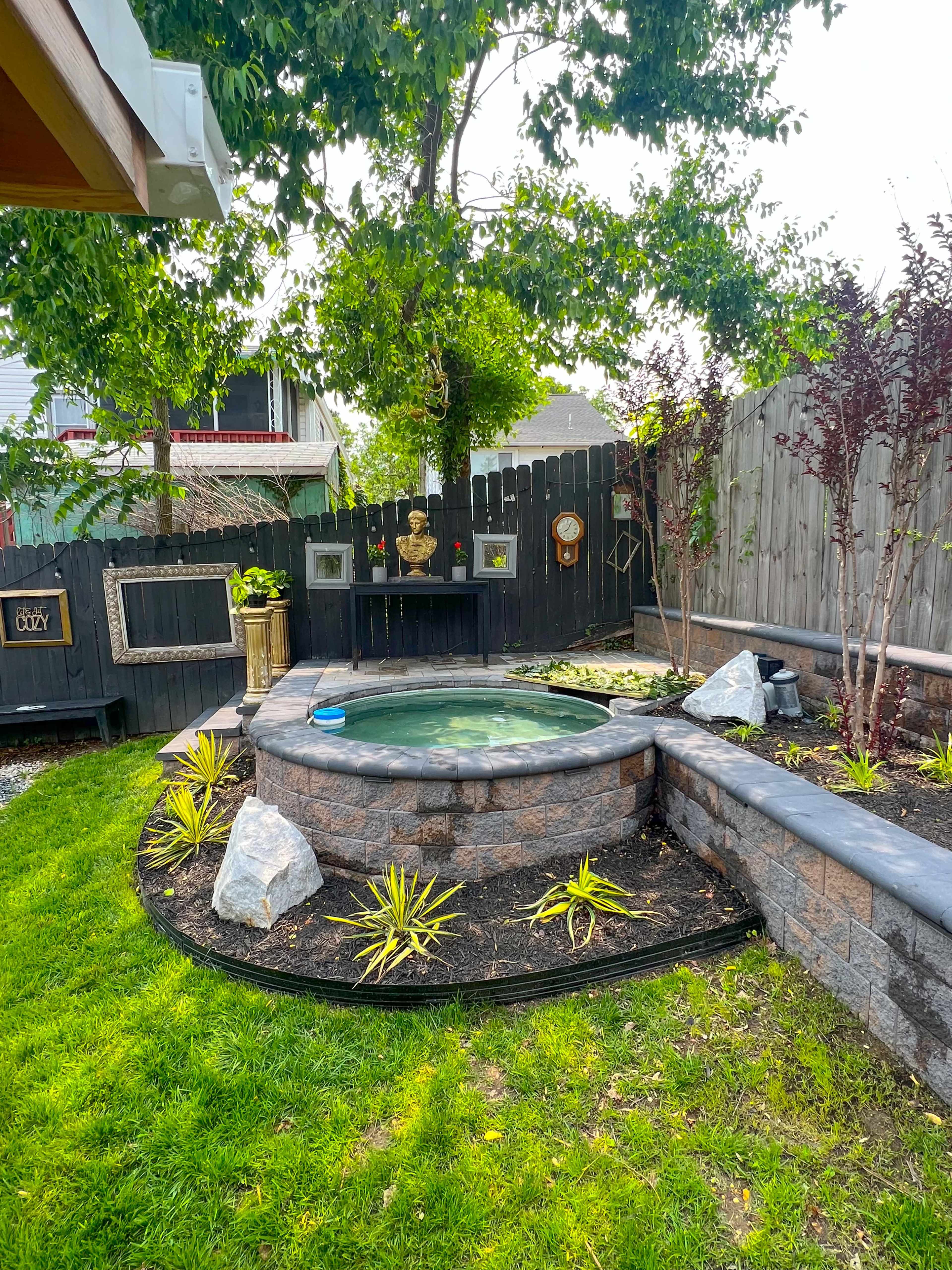 A landscaped backyard featuring a circular hot tub surrounded by stone walls, greenery, and decorative plants, with a wooden fence and various garden elements in the background.