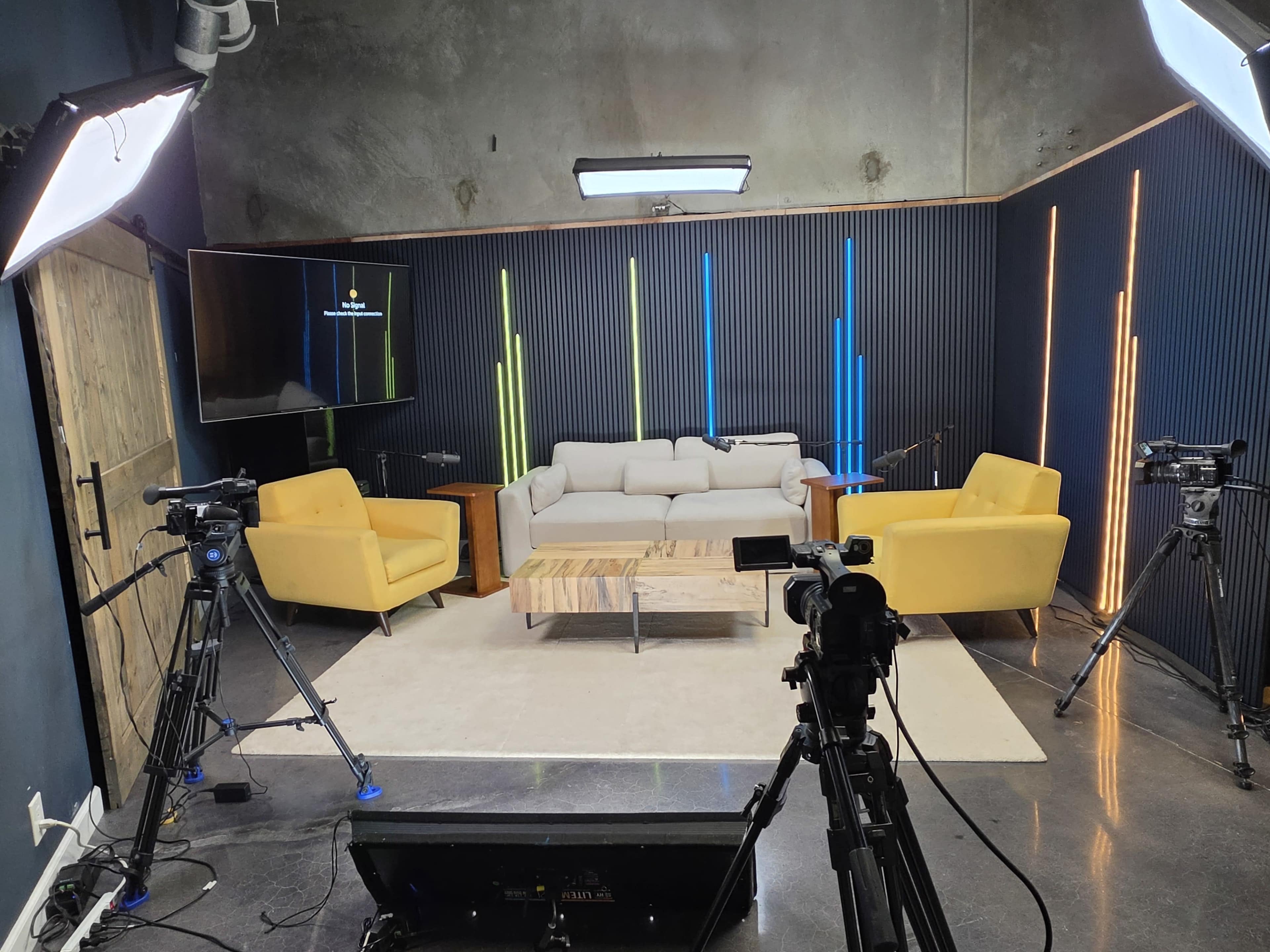 The image shows a modern studio setup with a sofa, two yellow chairs, a coffee table, and multiple cameras positioned on tripods.