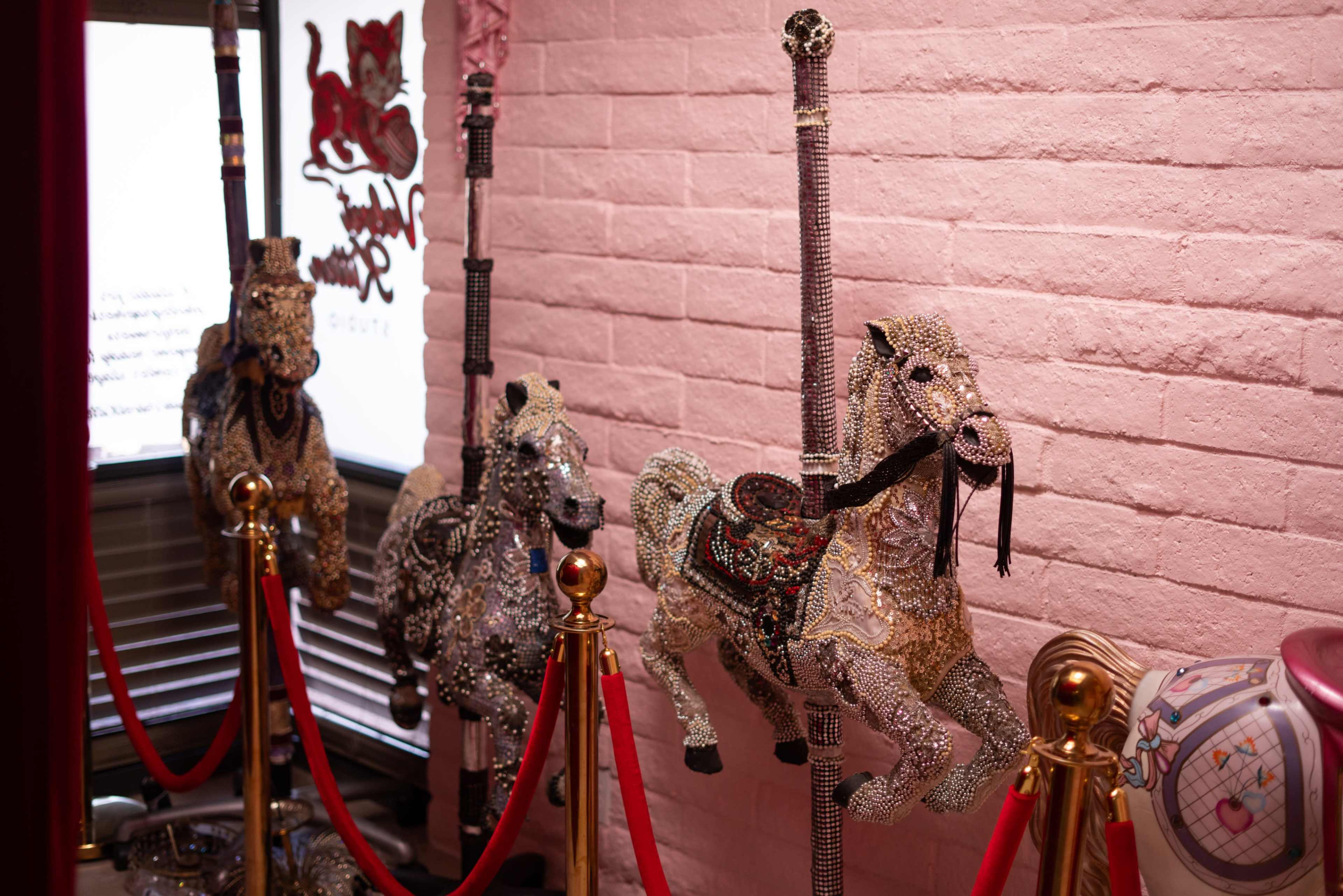 The image features three ornate carousel horses decorated with intricate designs, positioned behind a velvet rope in a pink-walled room.