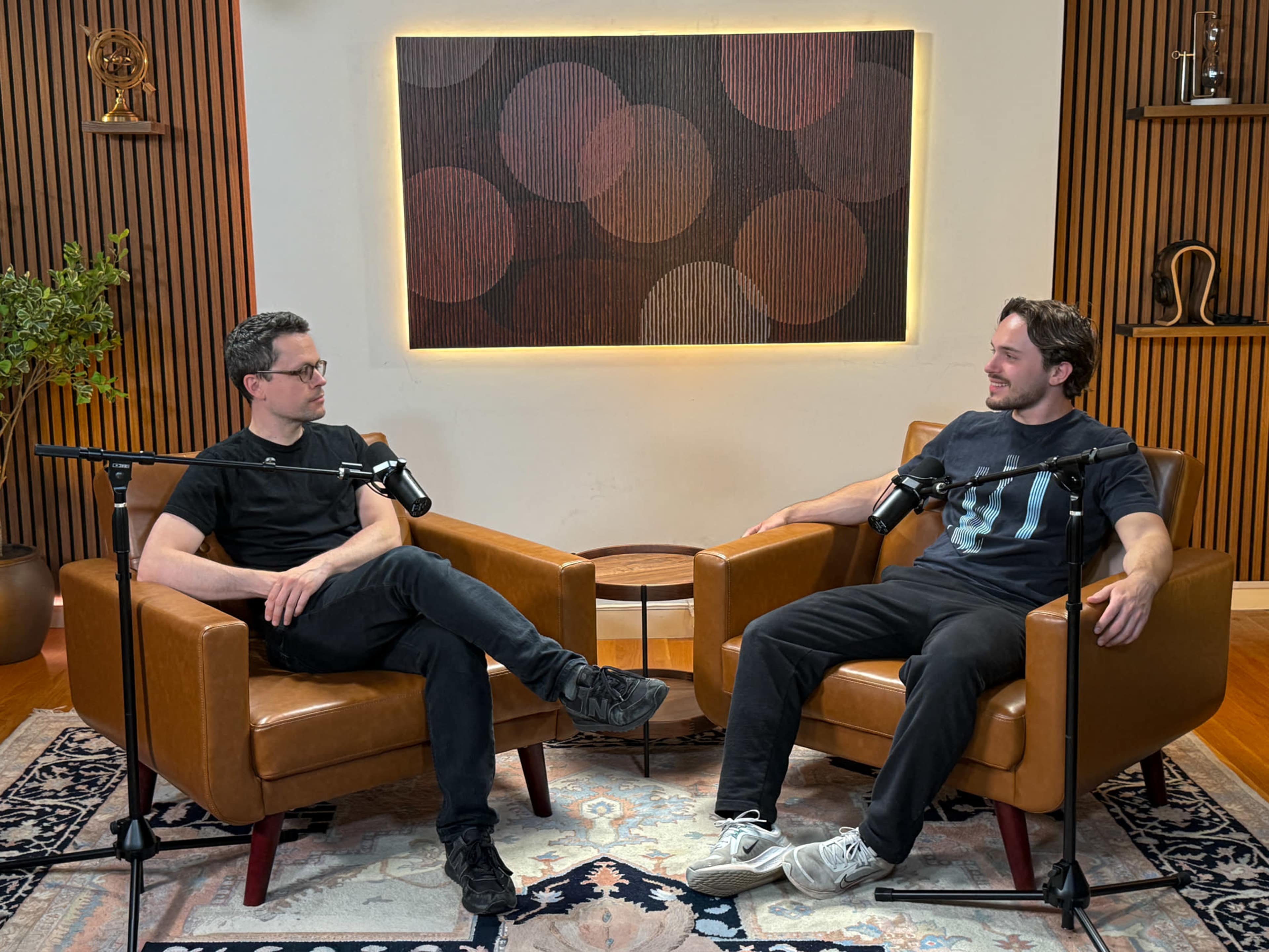 Two men sit in brown armchairs in a well-lit room, equipped with microphones, engaging in conversation.