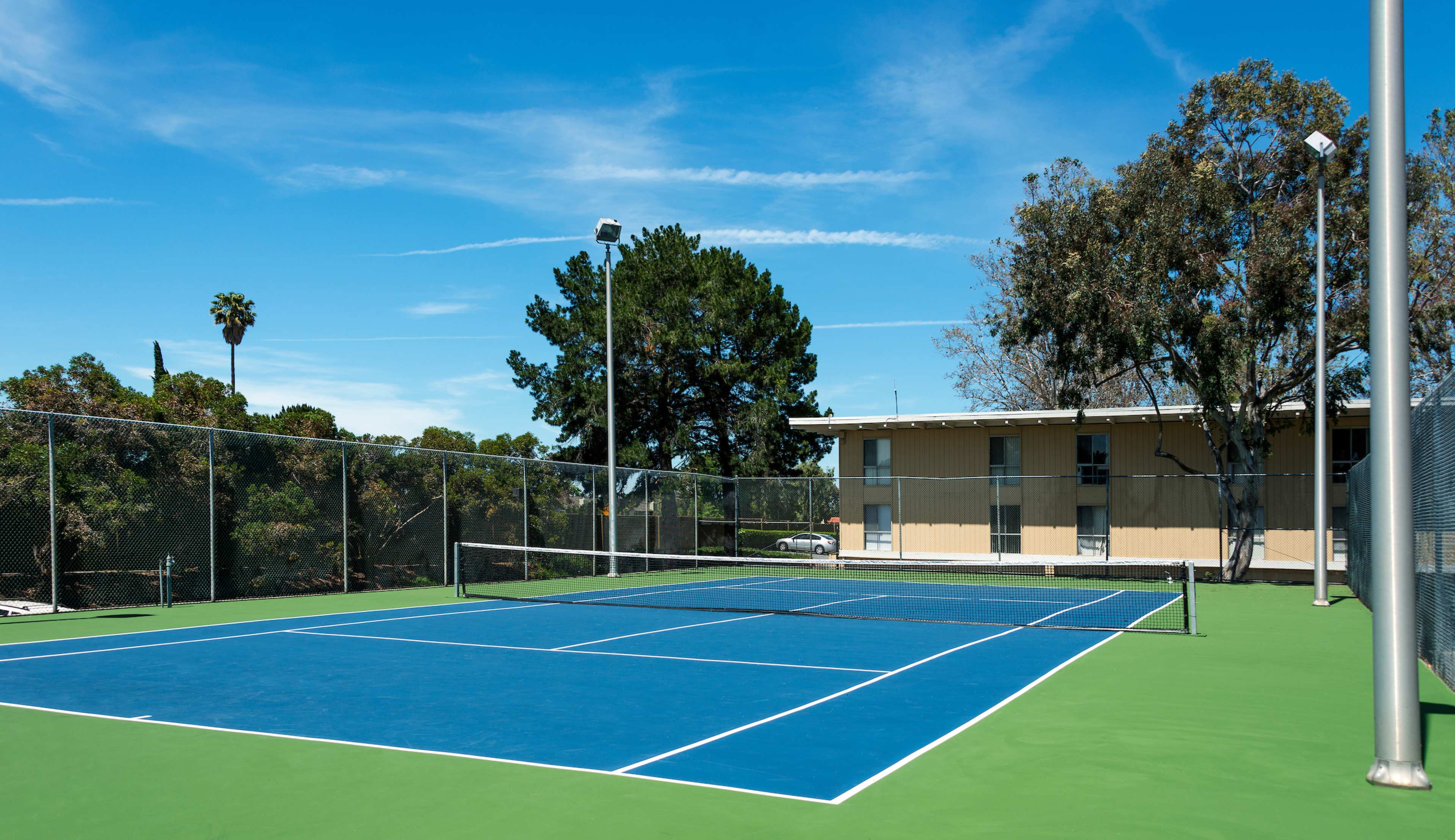 A bright outdoor tennis court with a green surface and blue lines is bordered by trees and a building in the background.