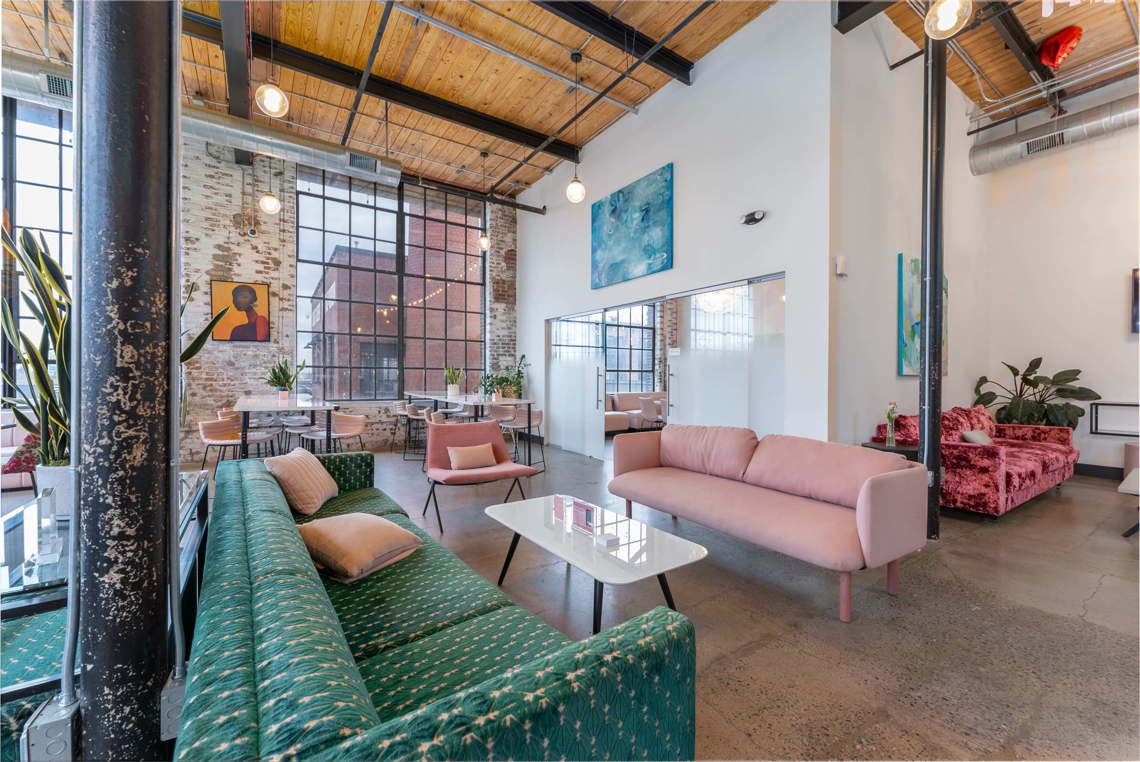 A modern lounge area with a green patterned sofa, a pink sofa, and several tables, complemented by large windows and artwork on the walls.
