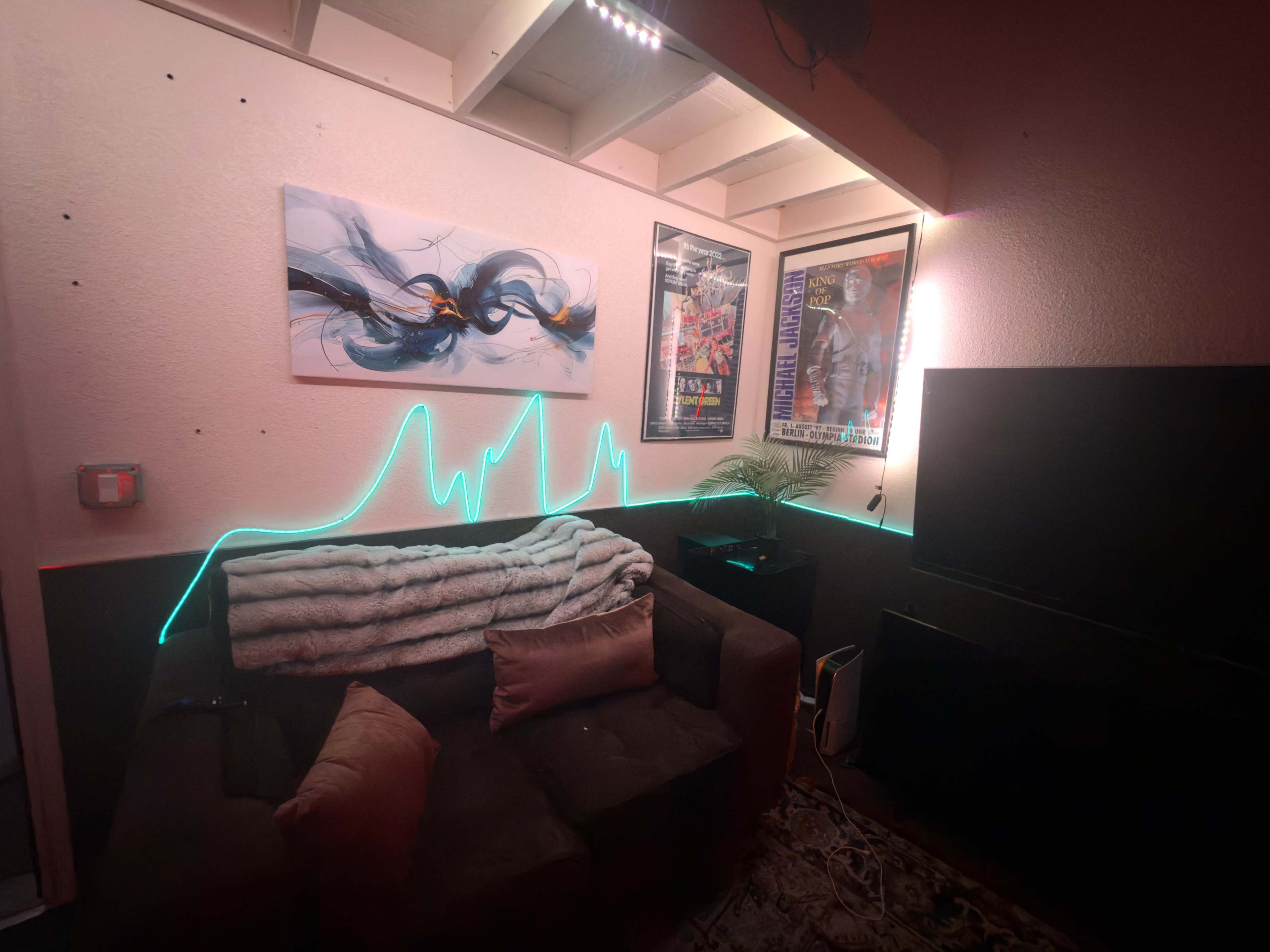 The image shows a corner of a living room featuring a couch, wall art, framed posters, a TV, and ambient LED lighting along the wall.