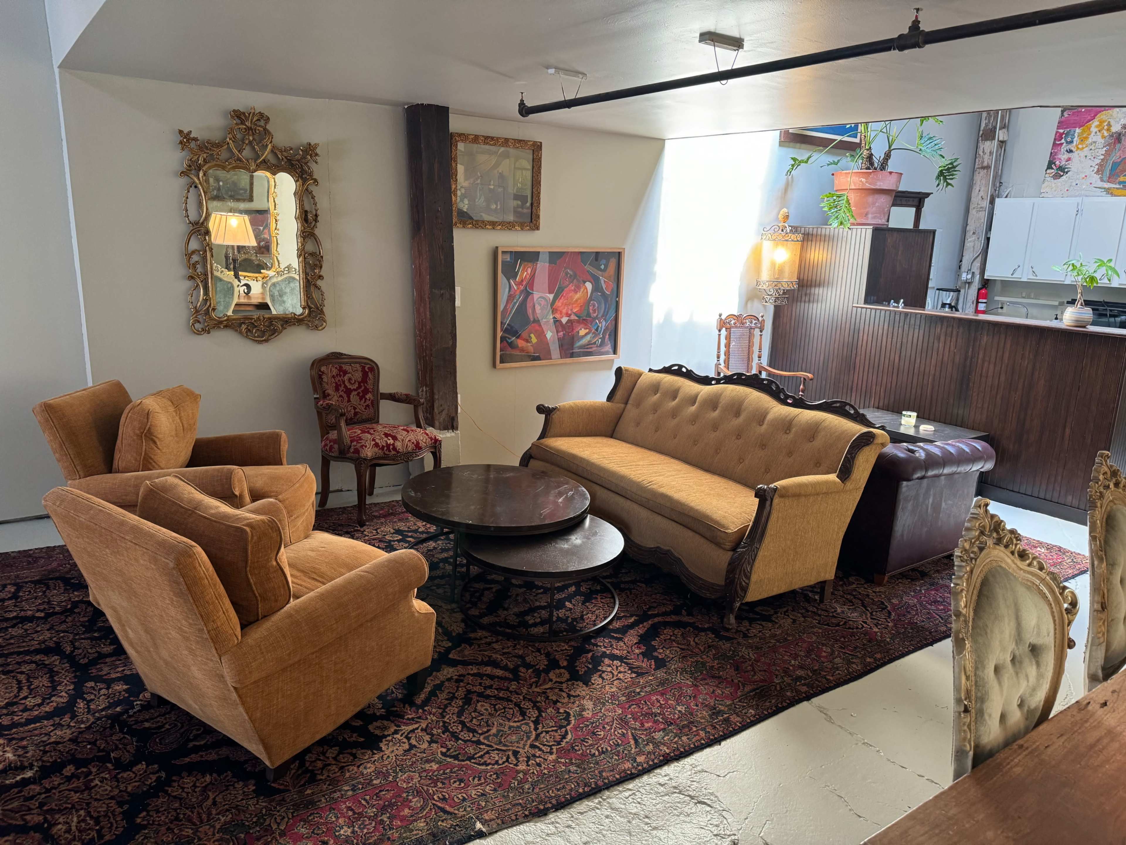 A cozy living room featuring a vintage-style sofa, two armchairs, a round coffee table, and an area rug, with a kitchenette visible in the background.