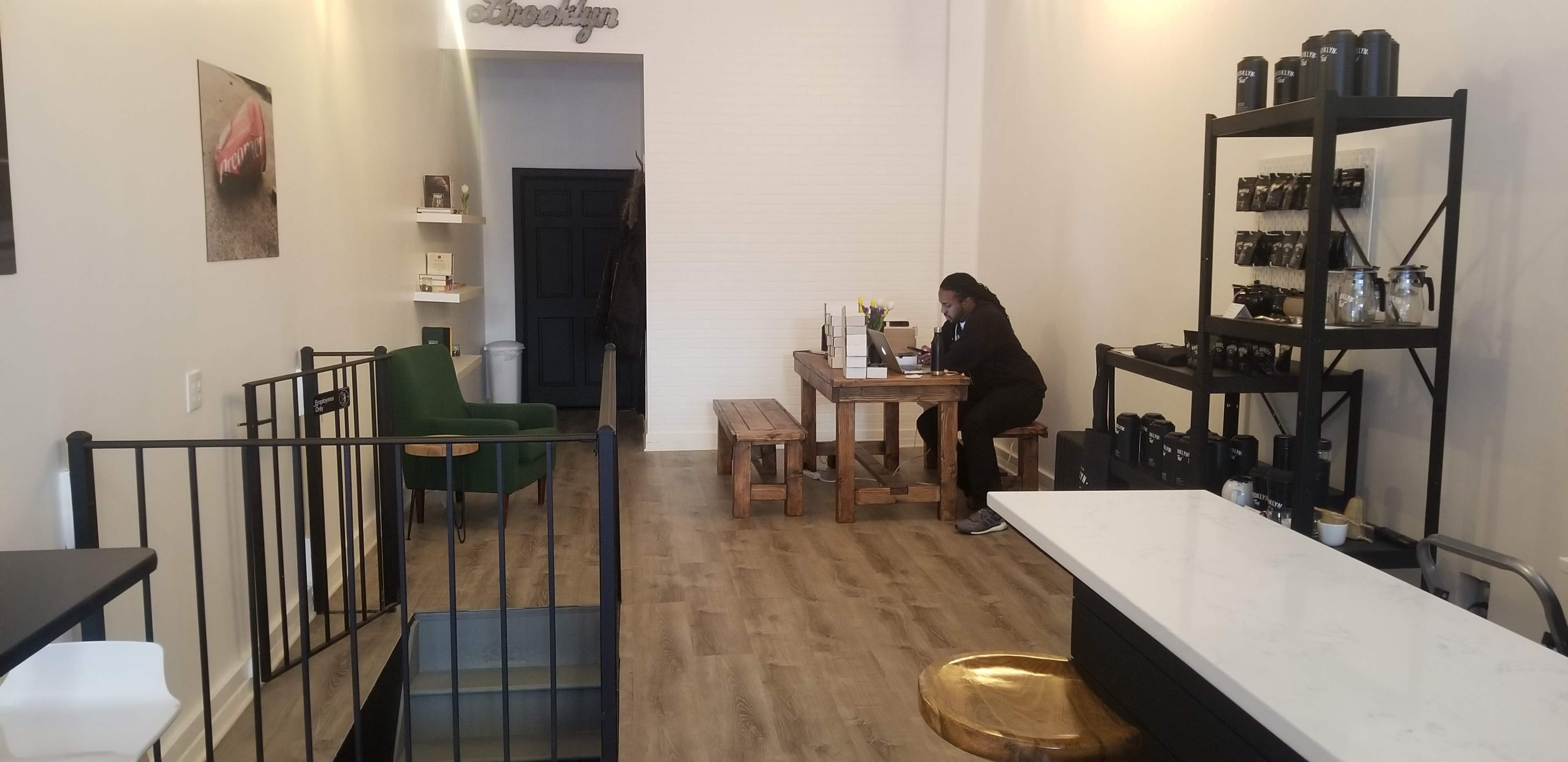 The image shows a minimalist café interior with a person sitting at a wooden table, surrounded by seating and shelves displaying various containers.