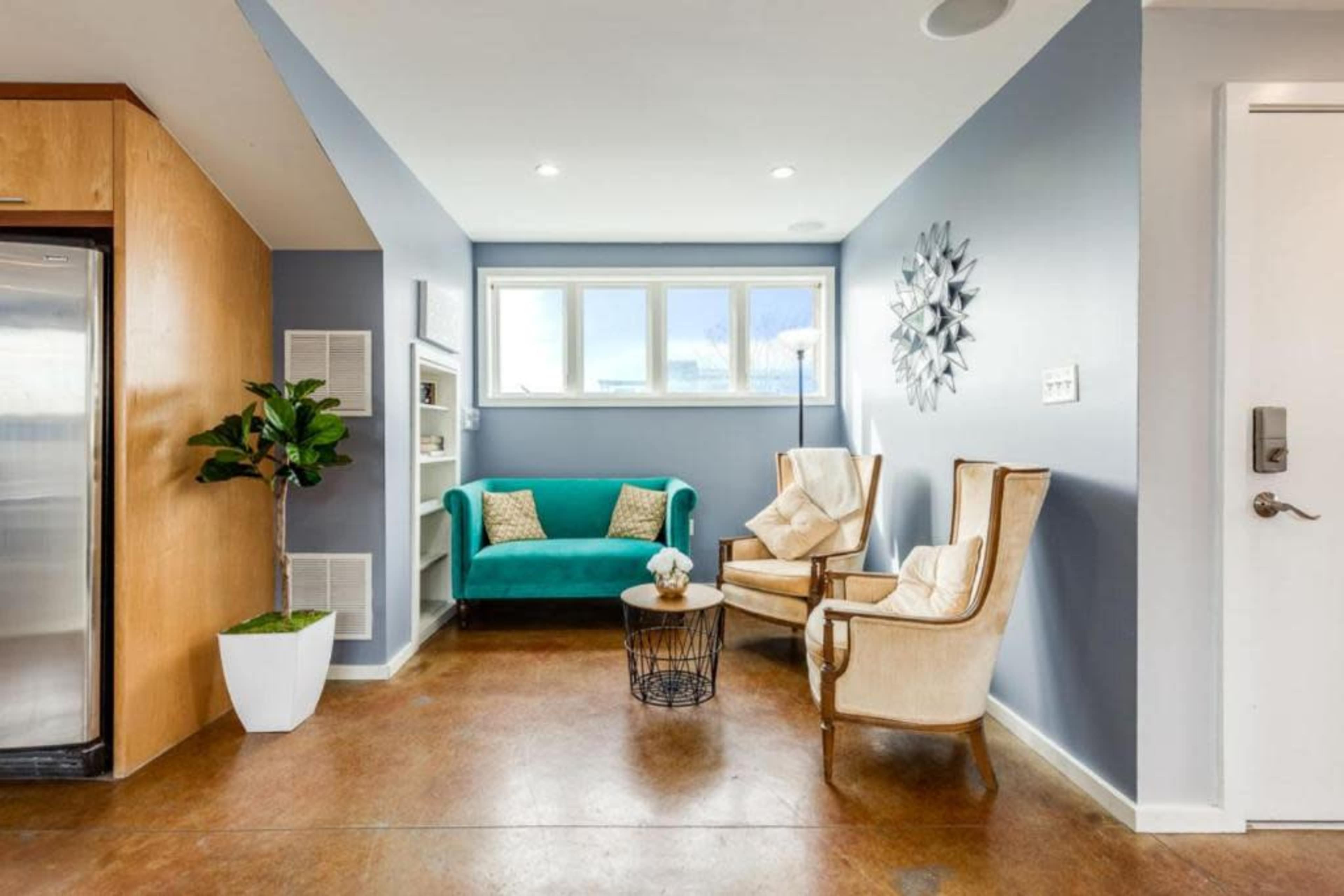 The image features a small, modern living area with a turquoise couch, two cream-colored armchairs, a coffee table, a potted plant, and large windows that let in natural light.