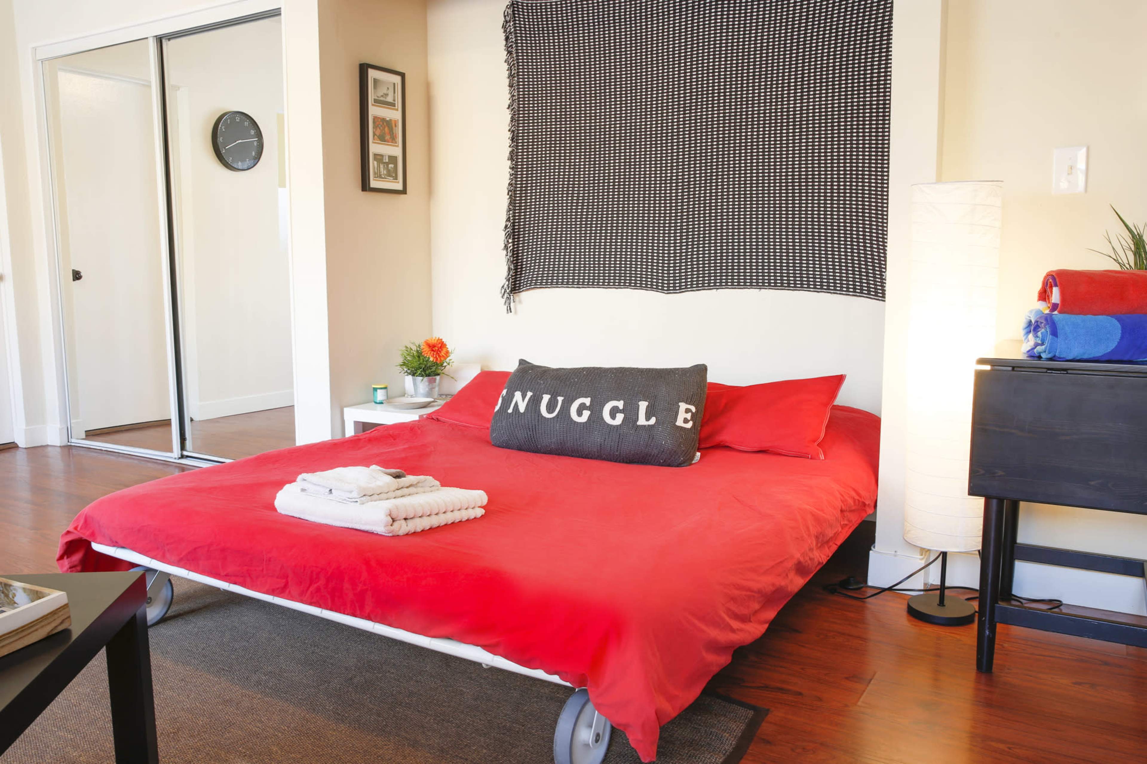 The image shows a neatly made bed with a red comforter and a large decorative pillow that reads "SNUGGLE," positioned against a light-colored wall with a black and white textile hanging above.