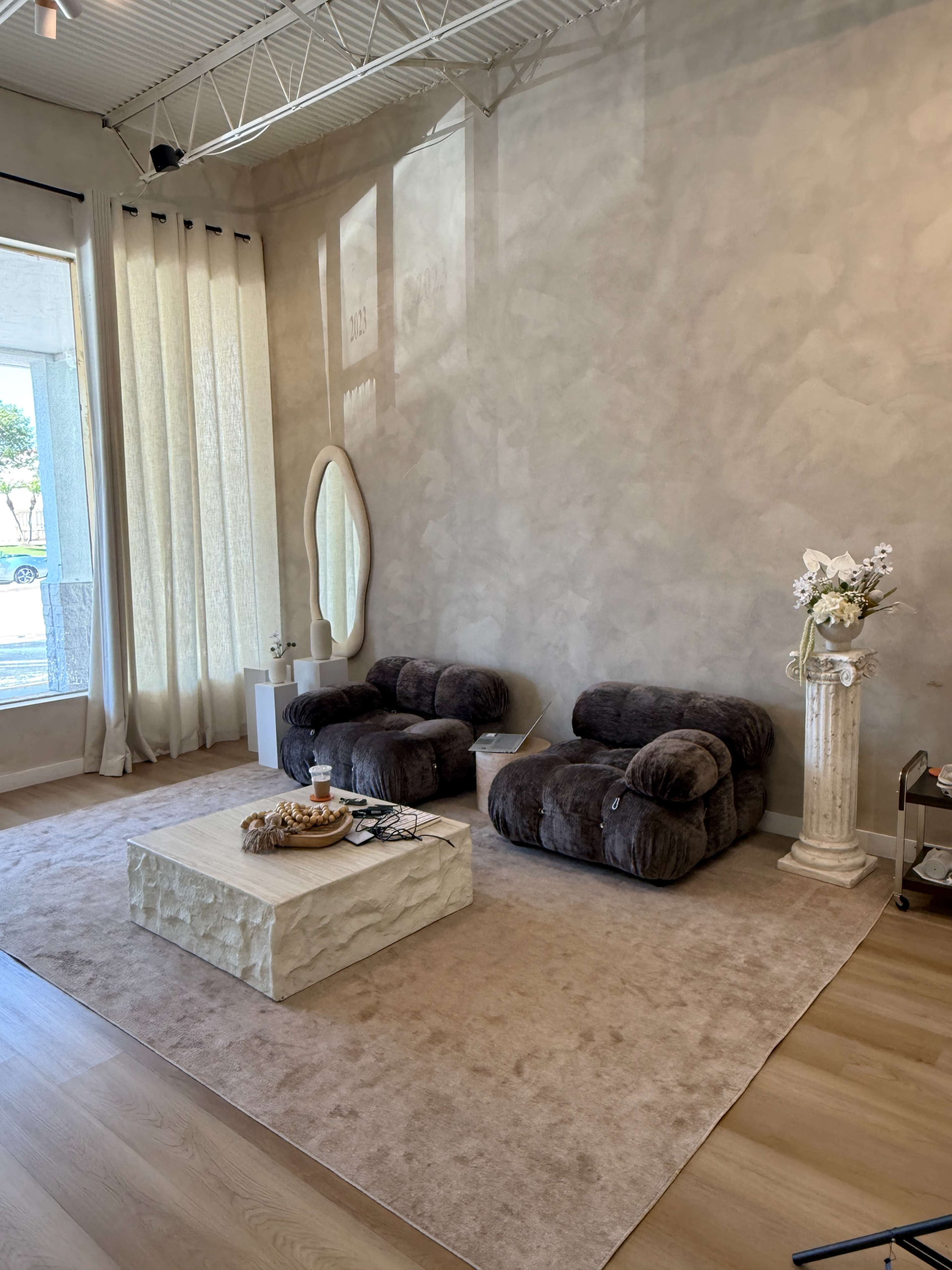 The image shows a modern interior with two plush chairs, a textured coffee table, and large windows allowing natural light to fill the space.