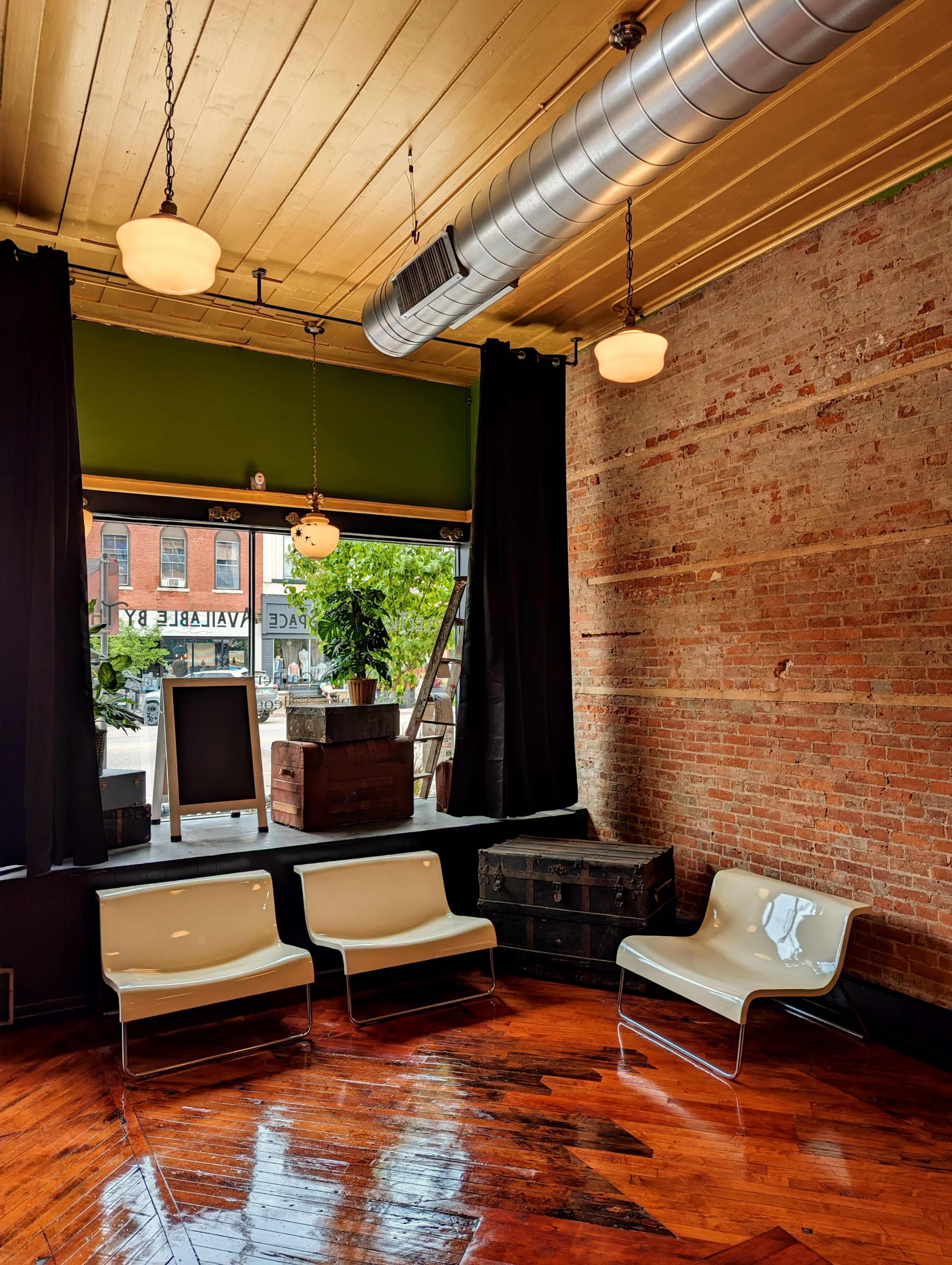 A minimalist interior of a café or lounge with three white chairs, a large window, and exposed brick walls.
