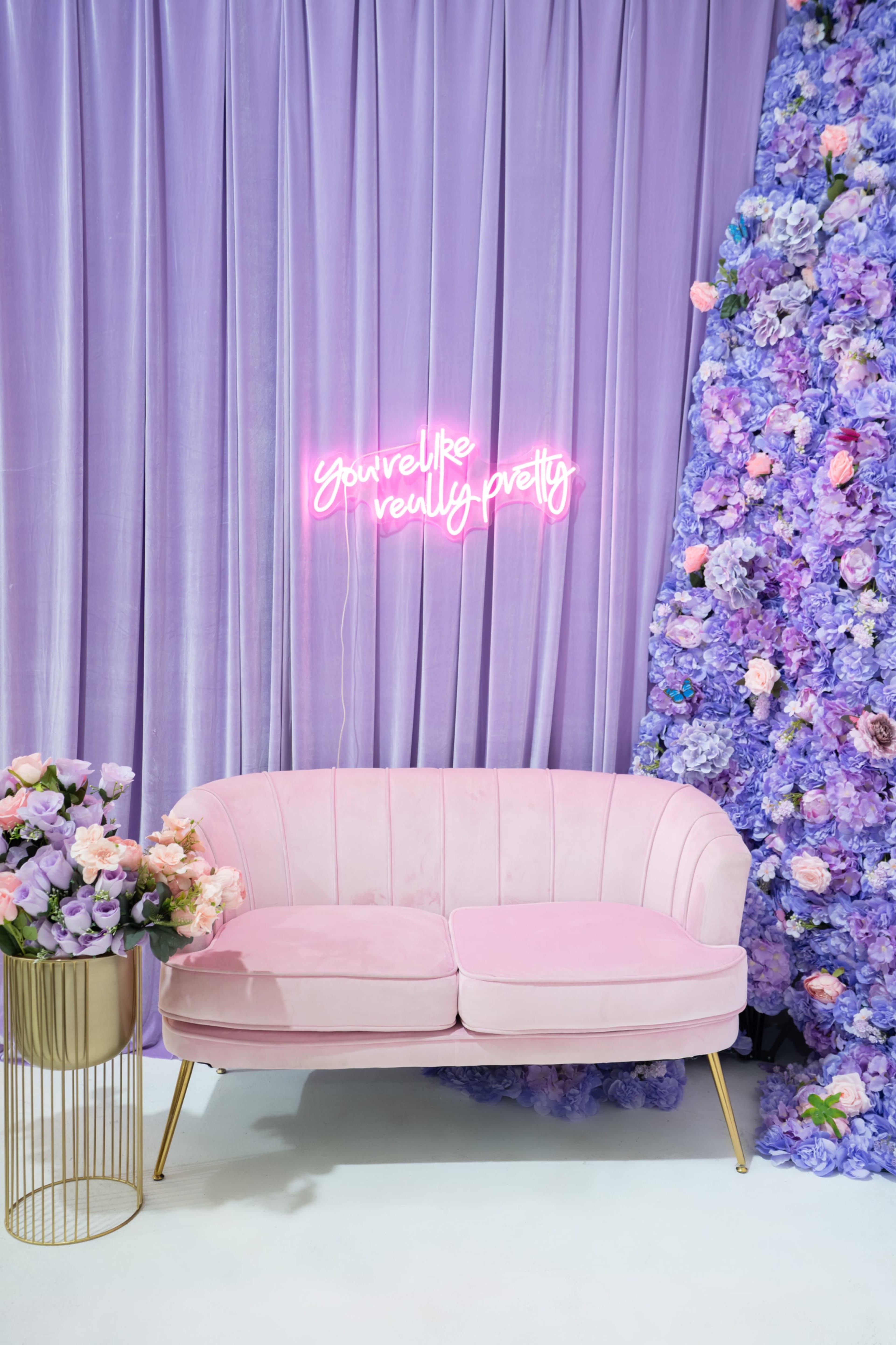 A pink velvet couch with gold legs sits in front of a lavender curtain and a vibrant floral arrangement, while a neon sign overhead reads "You're like really pretty."