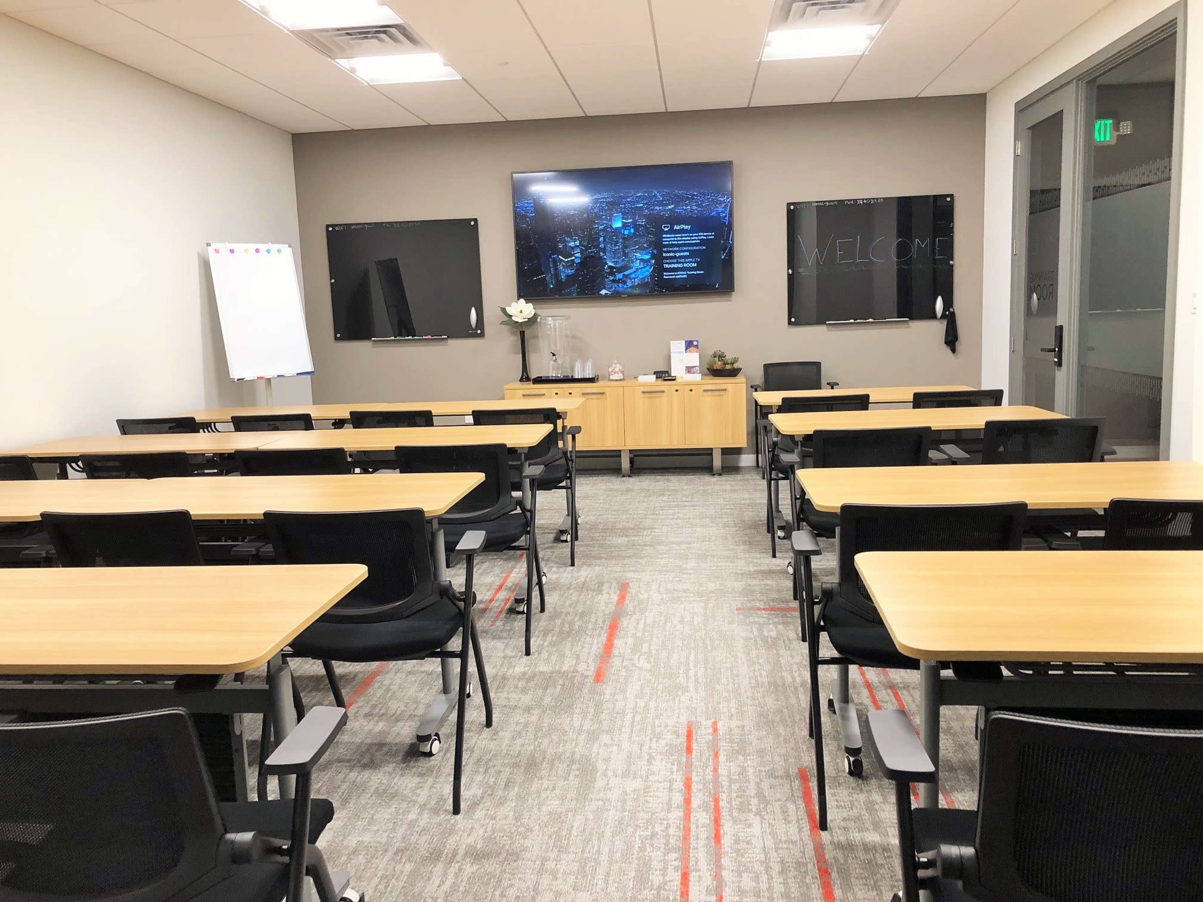 A modern meeting room is set up with several rectangular tables and chairs arranged facing a wall-mounted screen displaying a welcome message.