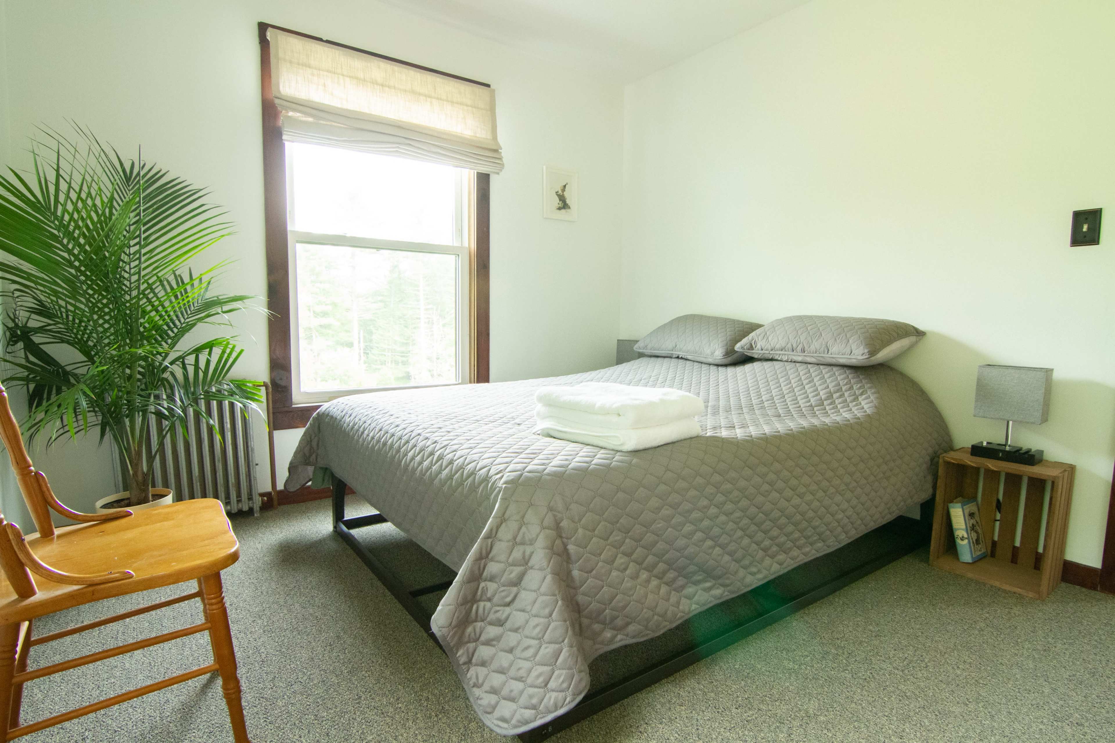 A neatly made bed with gray bedding is positioned in a bright room featuring a window, a potted plant, and a wooden chair.