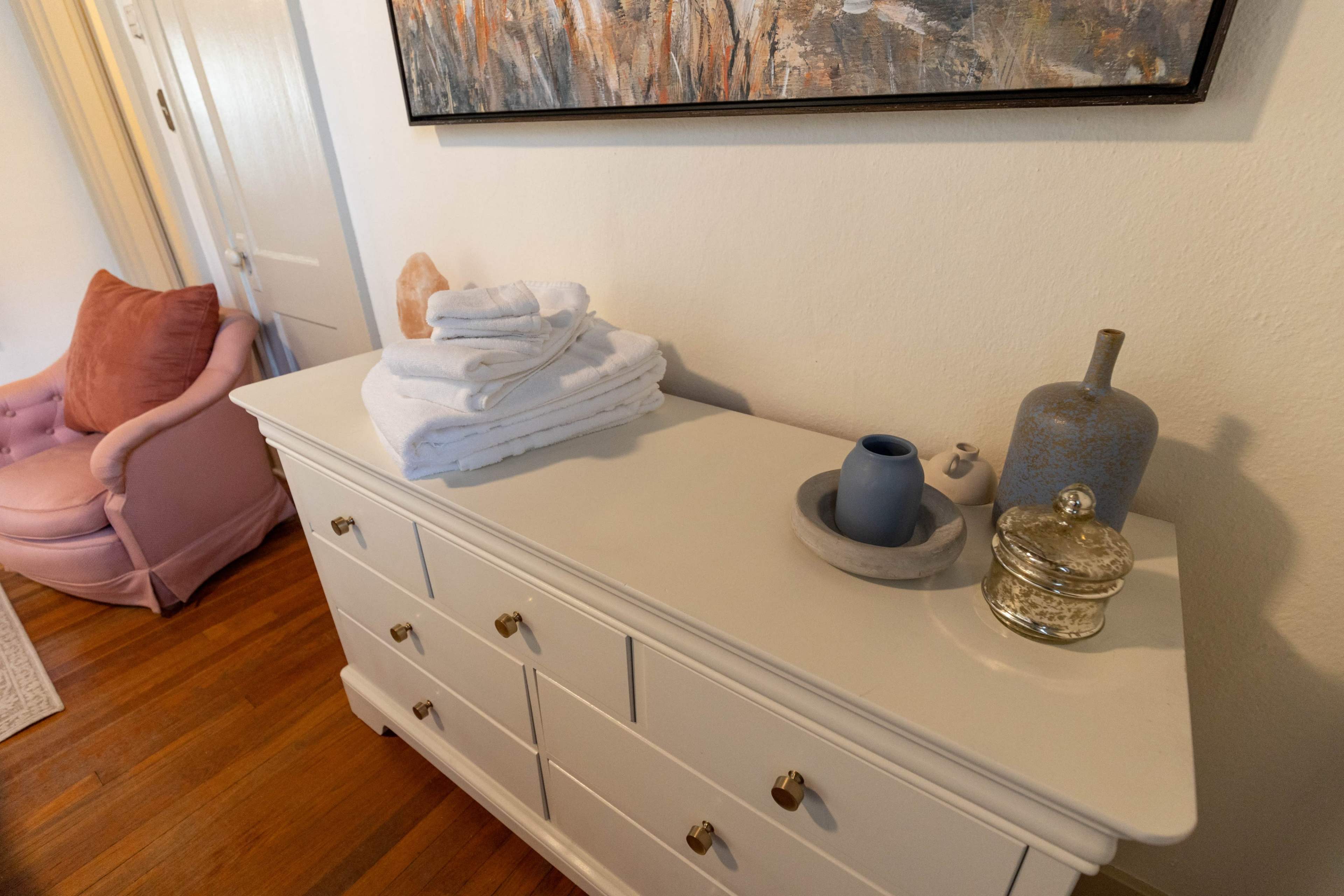 A neatly arranged dresser holds stacked towels, decorative items, and a small vase in a room with wooden flooring.