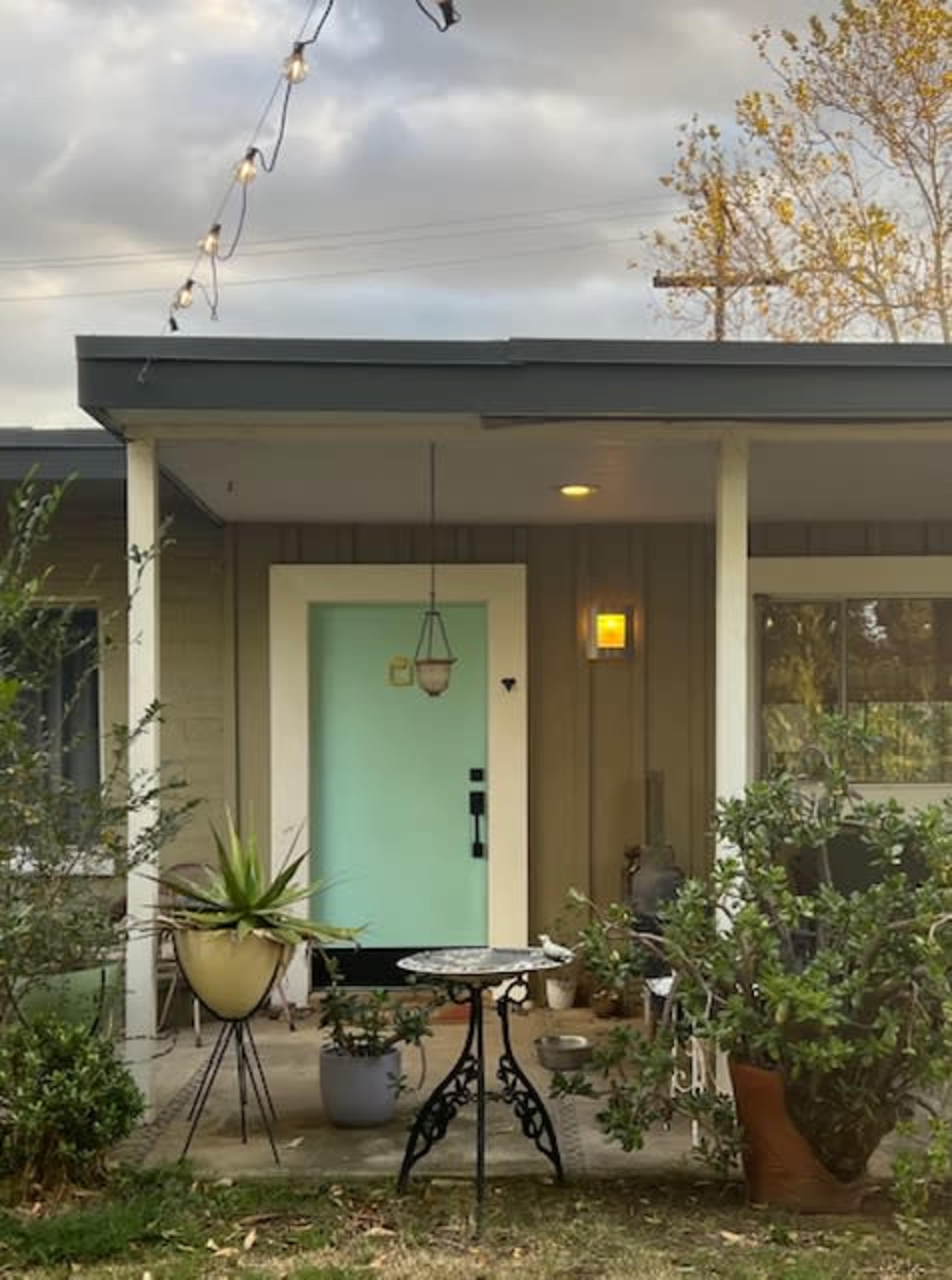 The image shows a front porch of a house with a light blue door, potted plants, and string lights overhead.