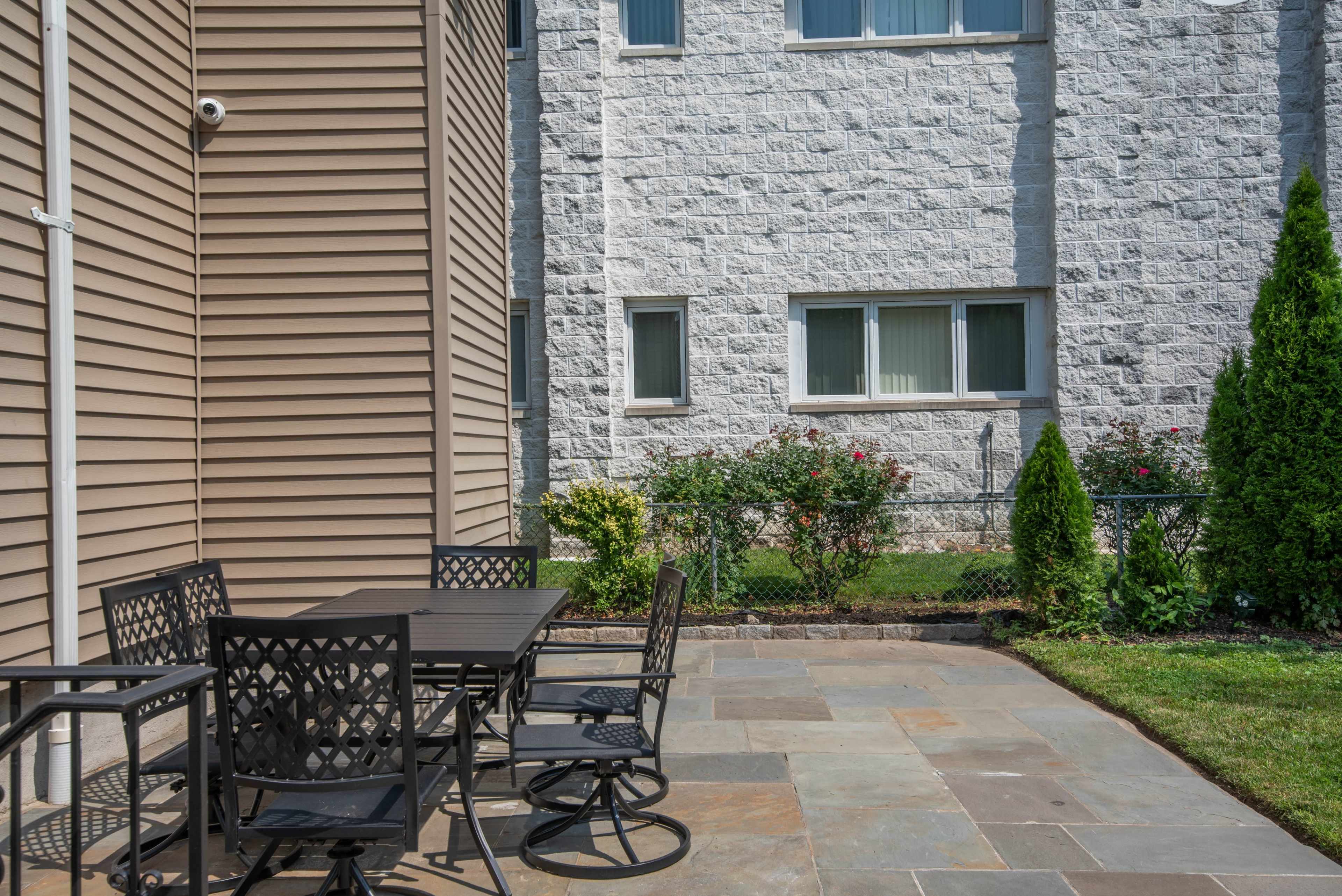 The image shows a stone patio with a black metal dining set situated next to a building with a textured gray wall and several windows.