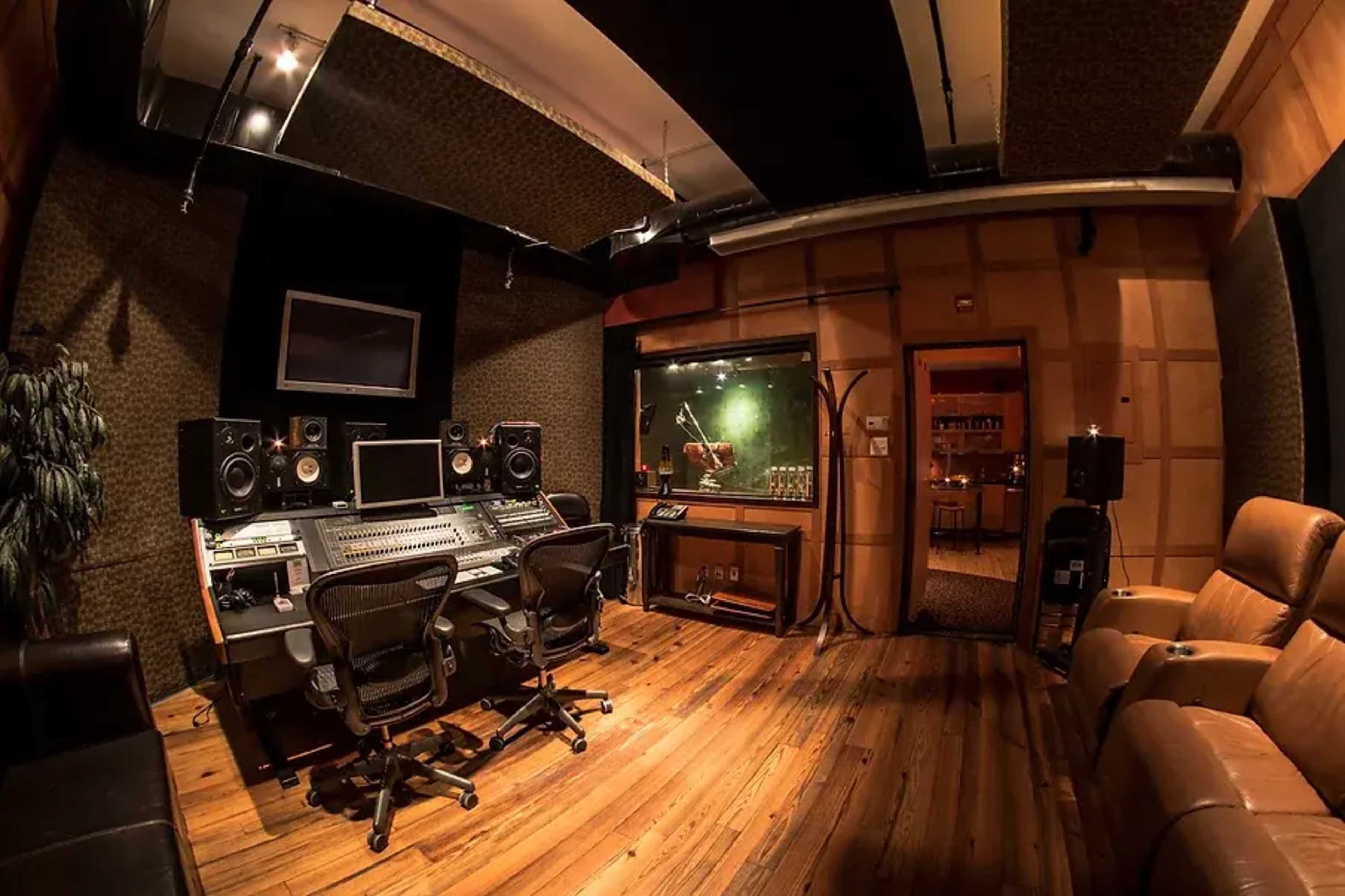 A soundproof recording studio featuring a large mixing console, two studio monitors, and a glass vocal booth alongside leather chairs and wooden flooring.