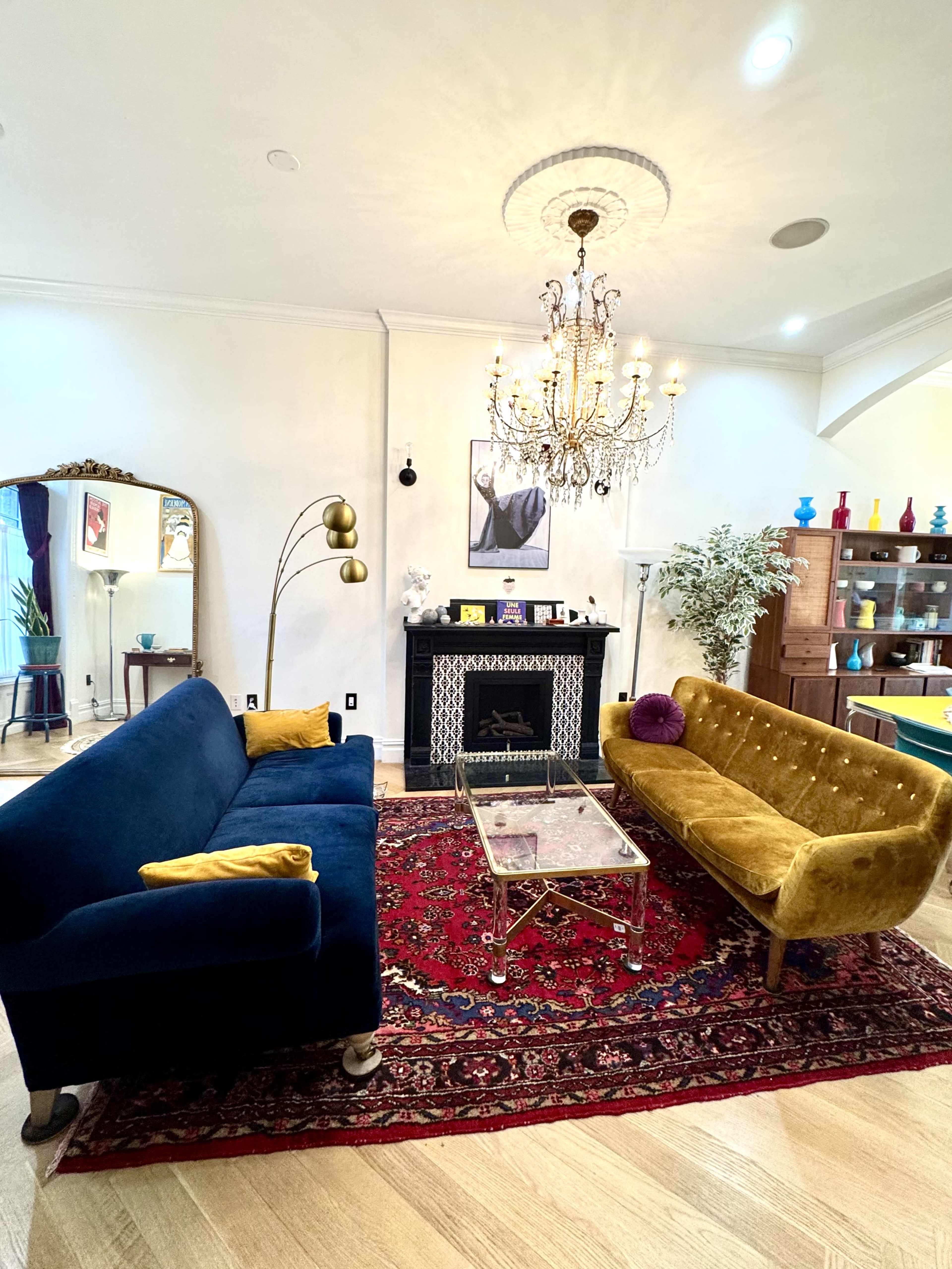 The image shows a stylish living room with a blue sofa, a yellow velvet couch, a glass coffee table on a red rug, and a chandelier above a decorative fireplace.