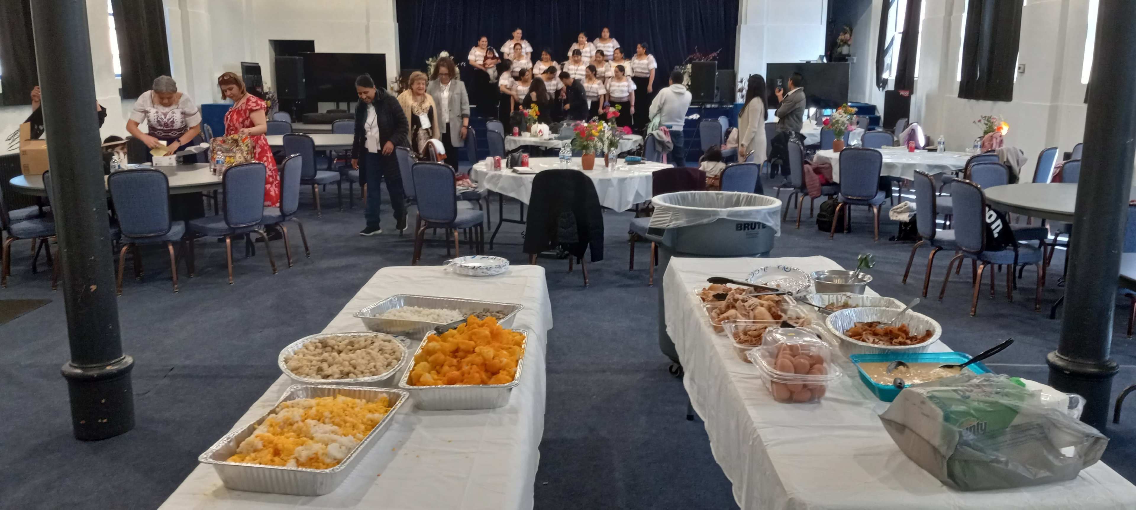 A dining area is set up with tables displaying food, while a group of people participates in a choir performance in the background.