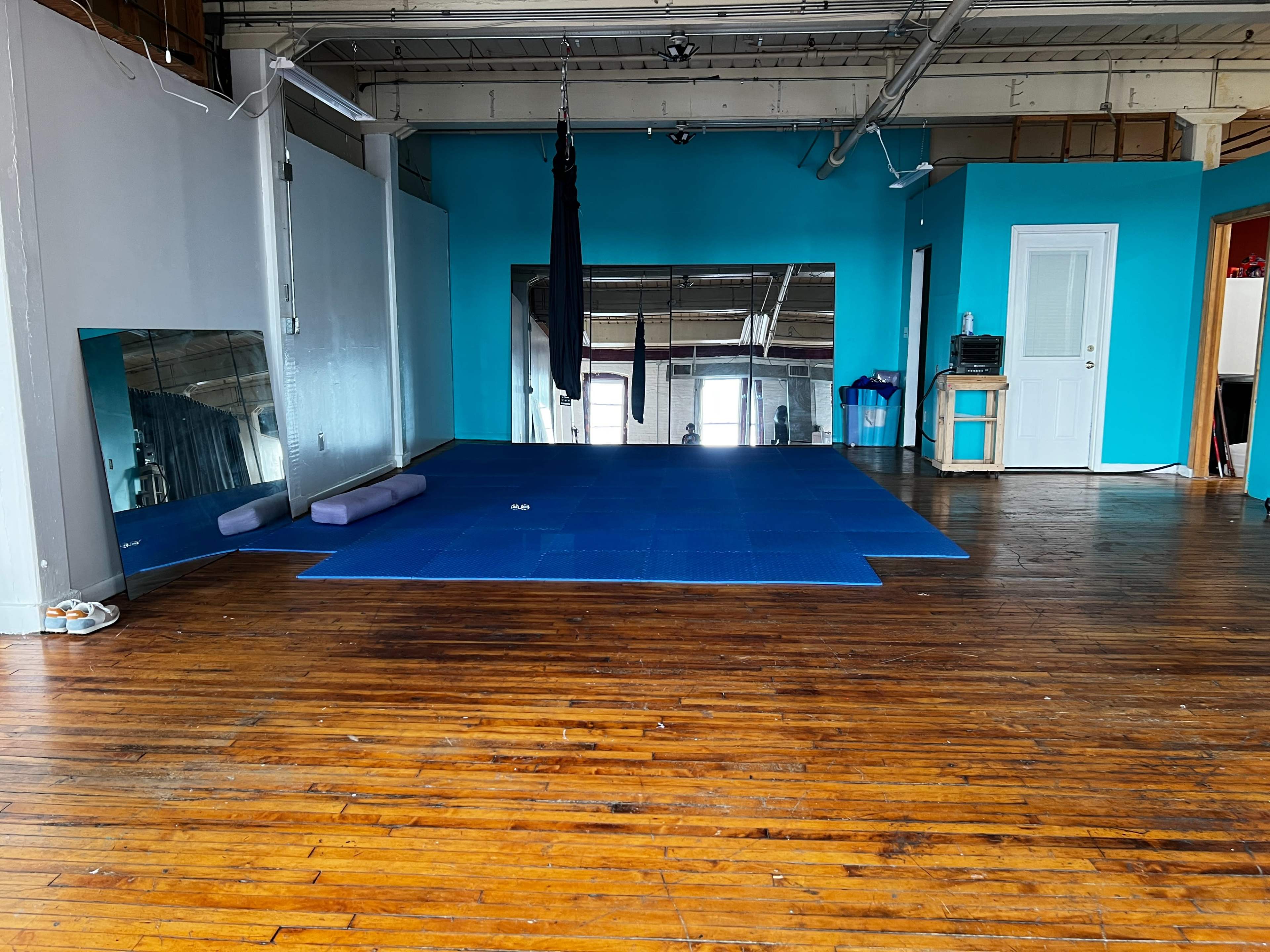 The image shows an empty studio space with blue walls, a blue exercise mat on the wooden floor, and large mirrors on one side.