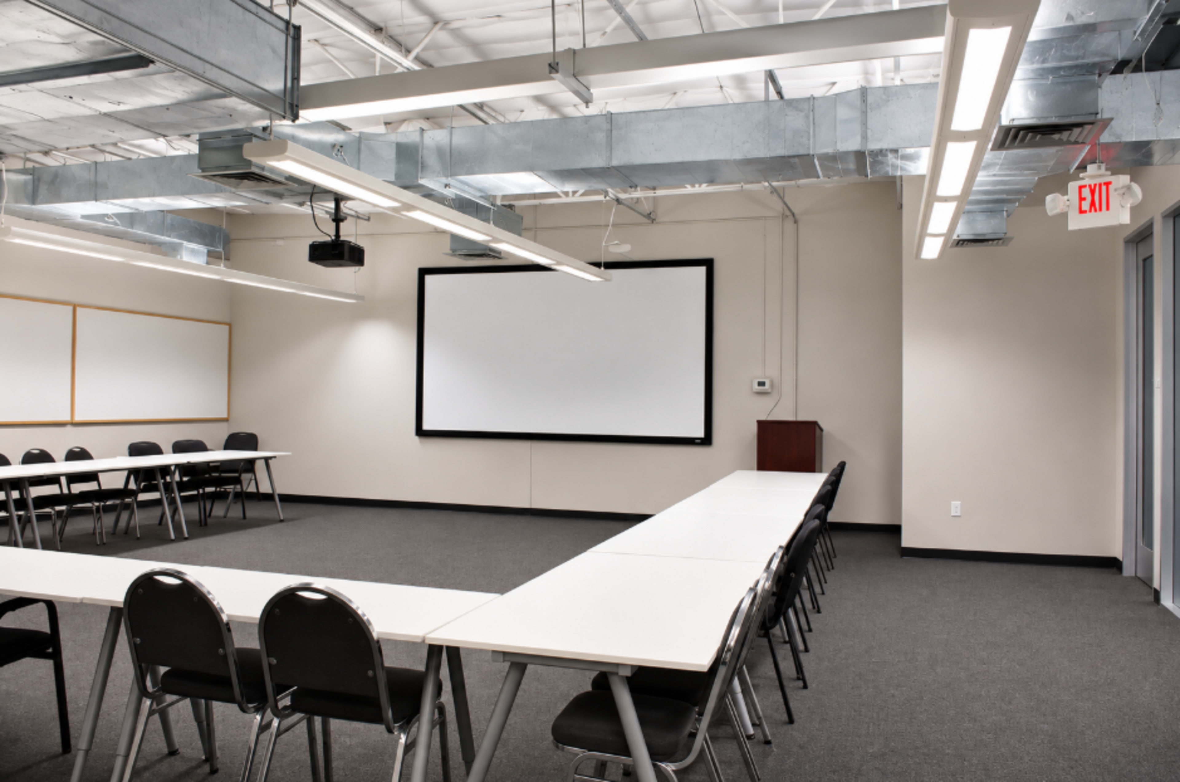 The image shows a modern conference room with a long table, black chairs, a blank projection screen on the wall, and exit signage.