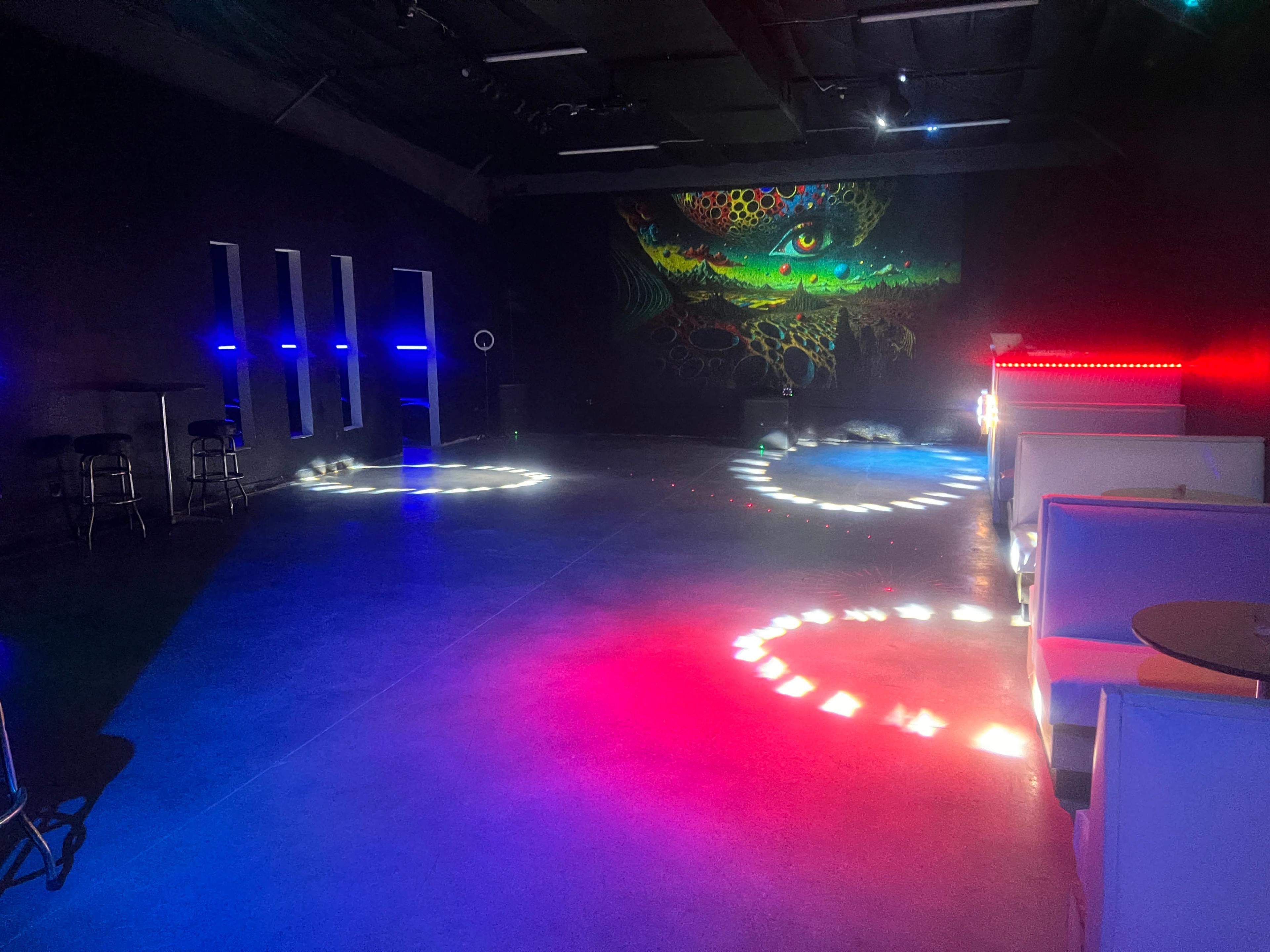 The image shows a dark, spacious venue with a colorful mural on the wall and various circular light patterns projected on the floor.