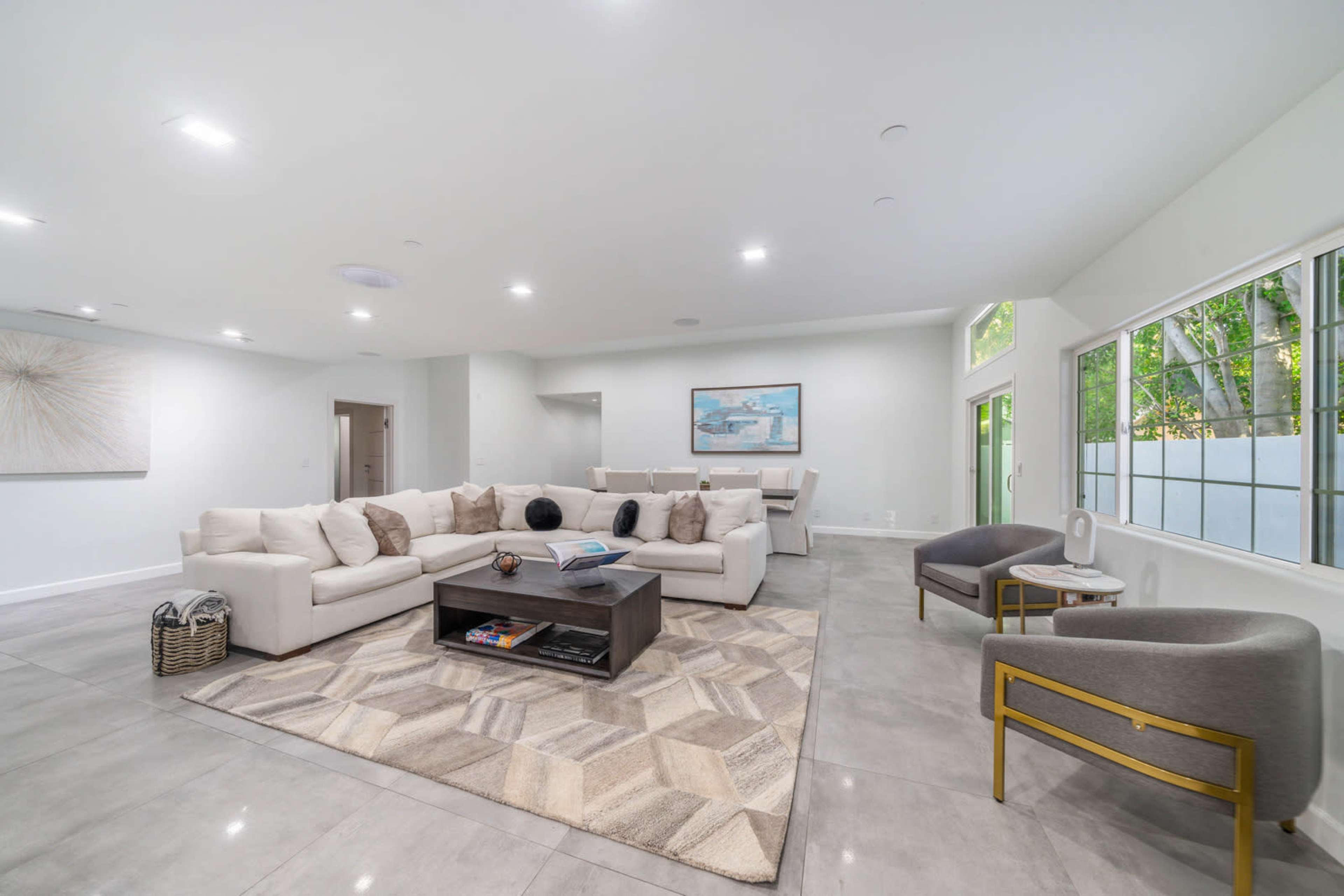 The image shows a spacious modern living room with a large sectional sofa, a coffee table, and natural light coming in through several windows.