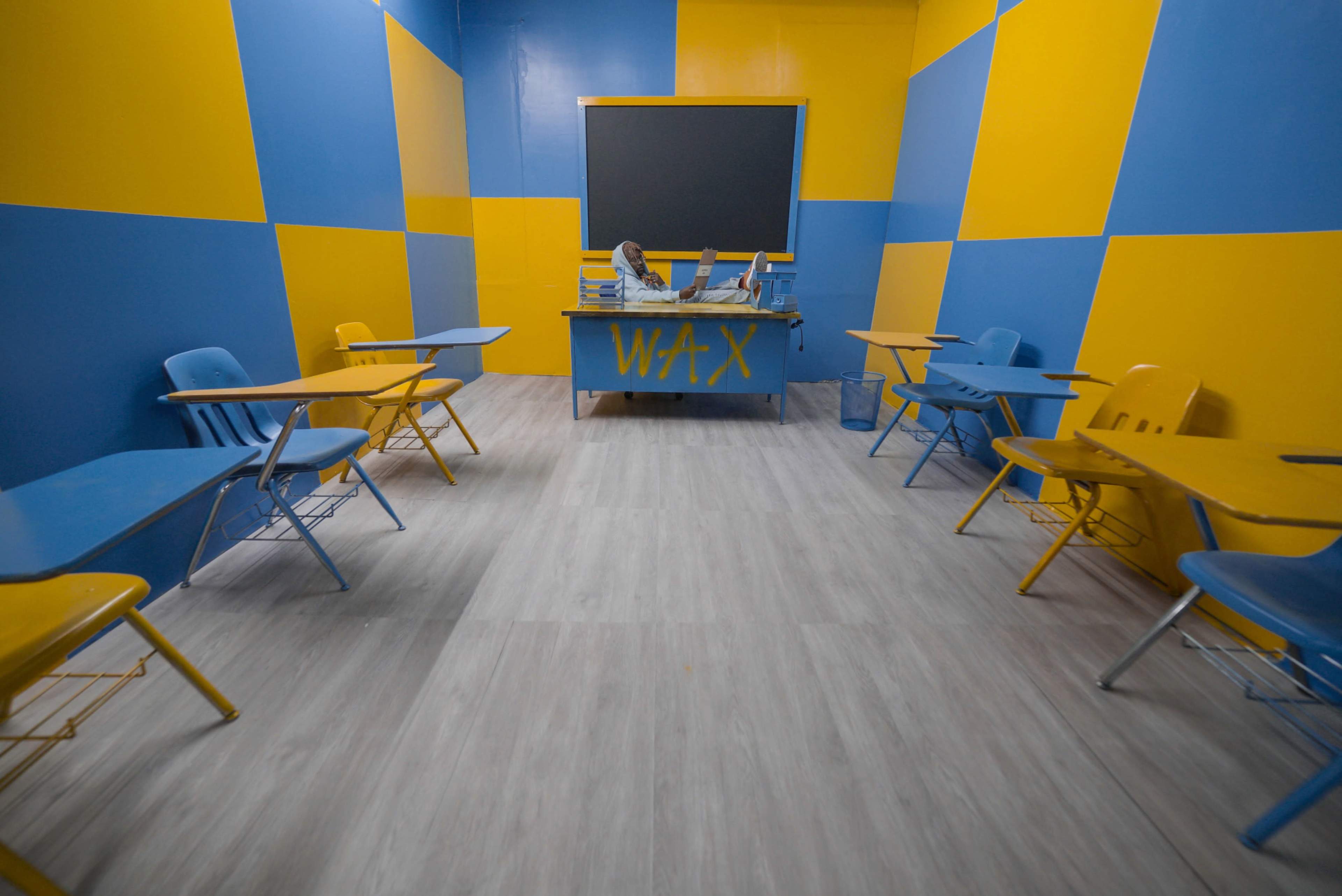 A classroom featuring blue and yellow walls, with a central desk labeled "WAX" and several empty blue and yellow desks arranged around the room.