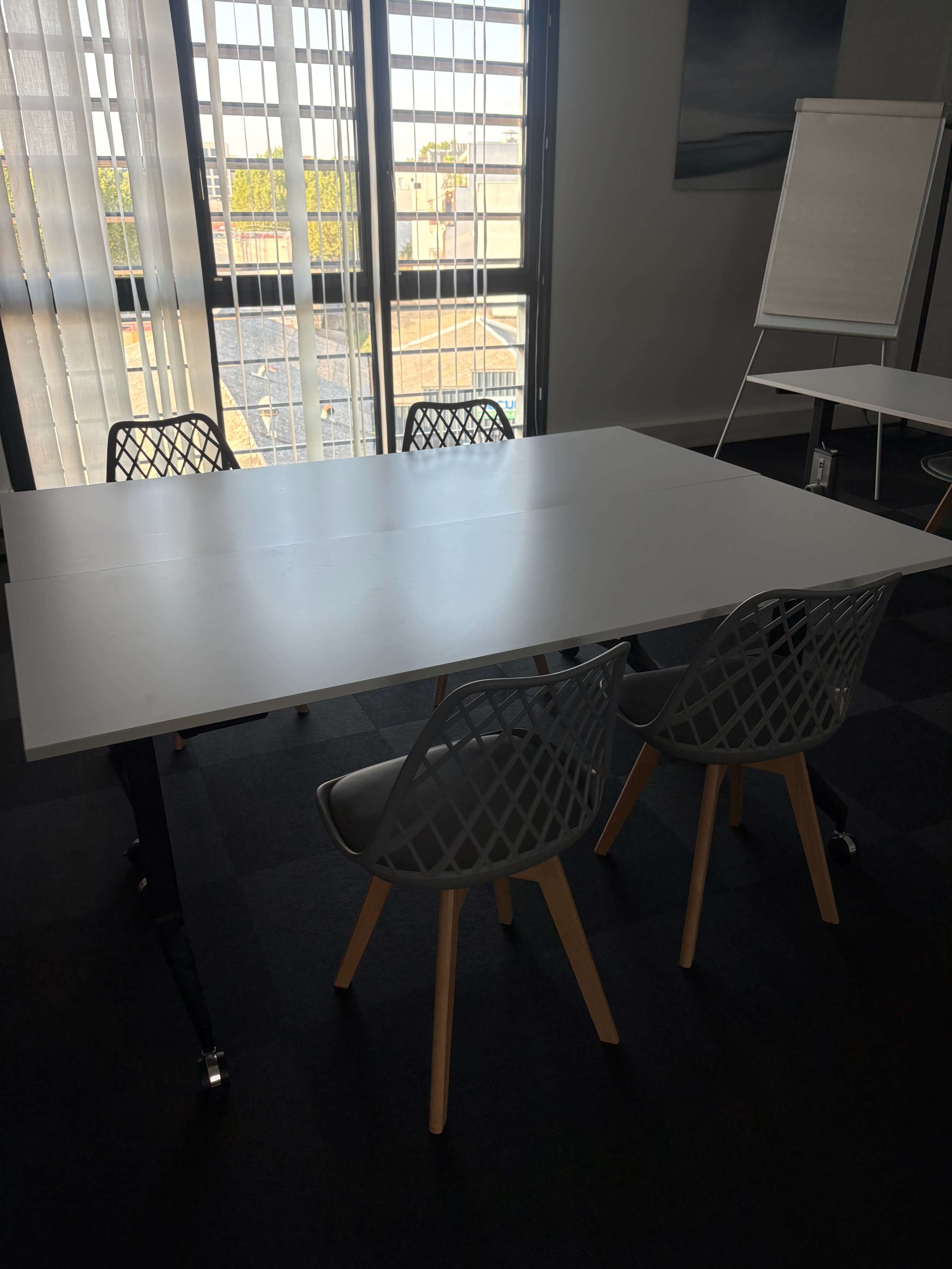 A modern meeting room features a white table surrounded by four gray chairs with wooden legs, near large windows with blinds.