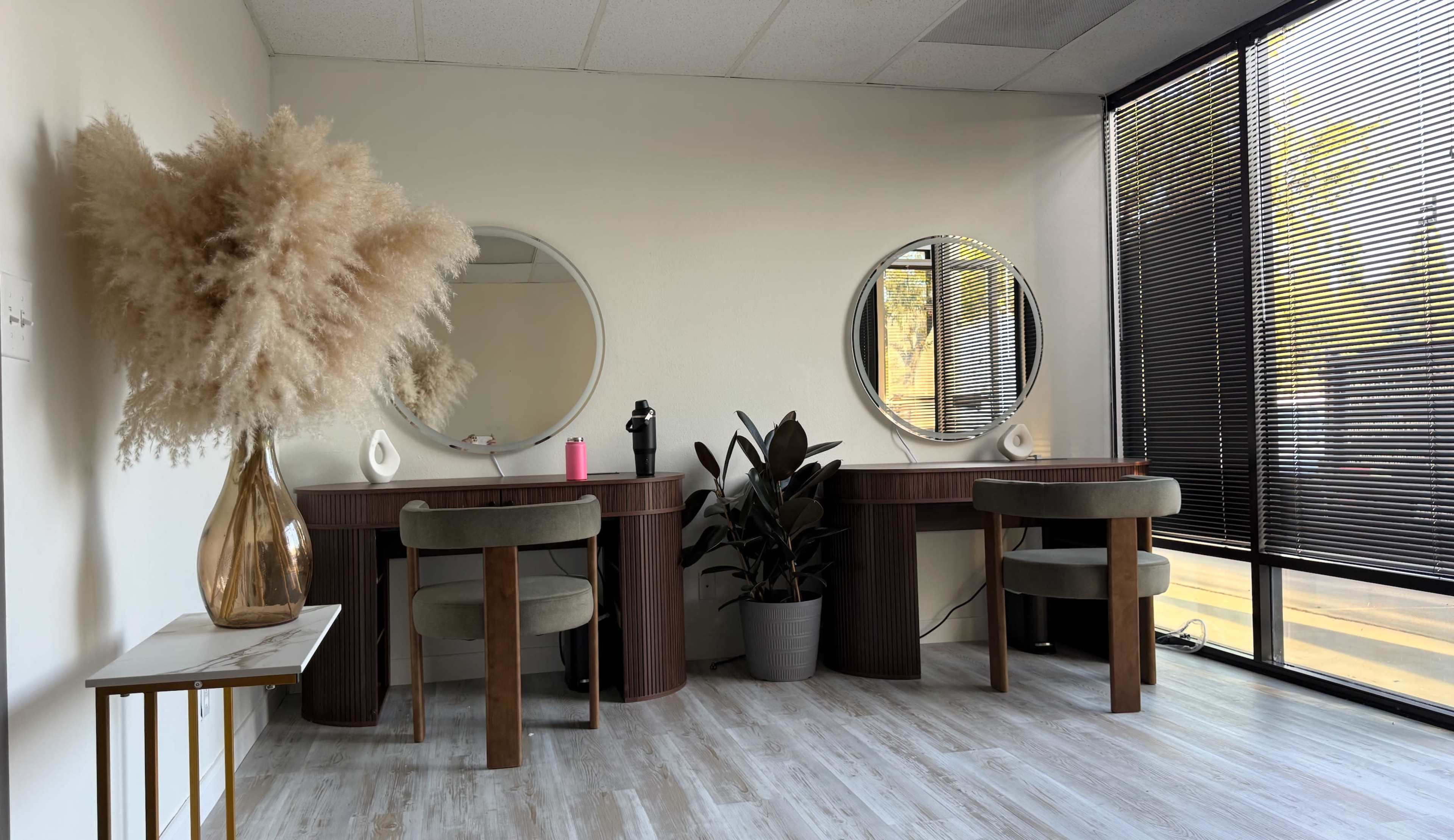 The image shows a modern beauty salon interior with two circular mirrors above elegant vanity tables, surrounded by a decorative plant and a large vase of dried pampas grass.