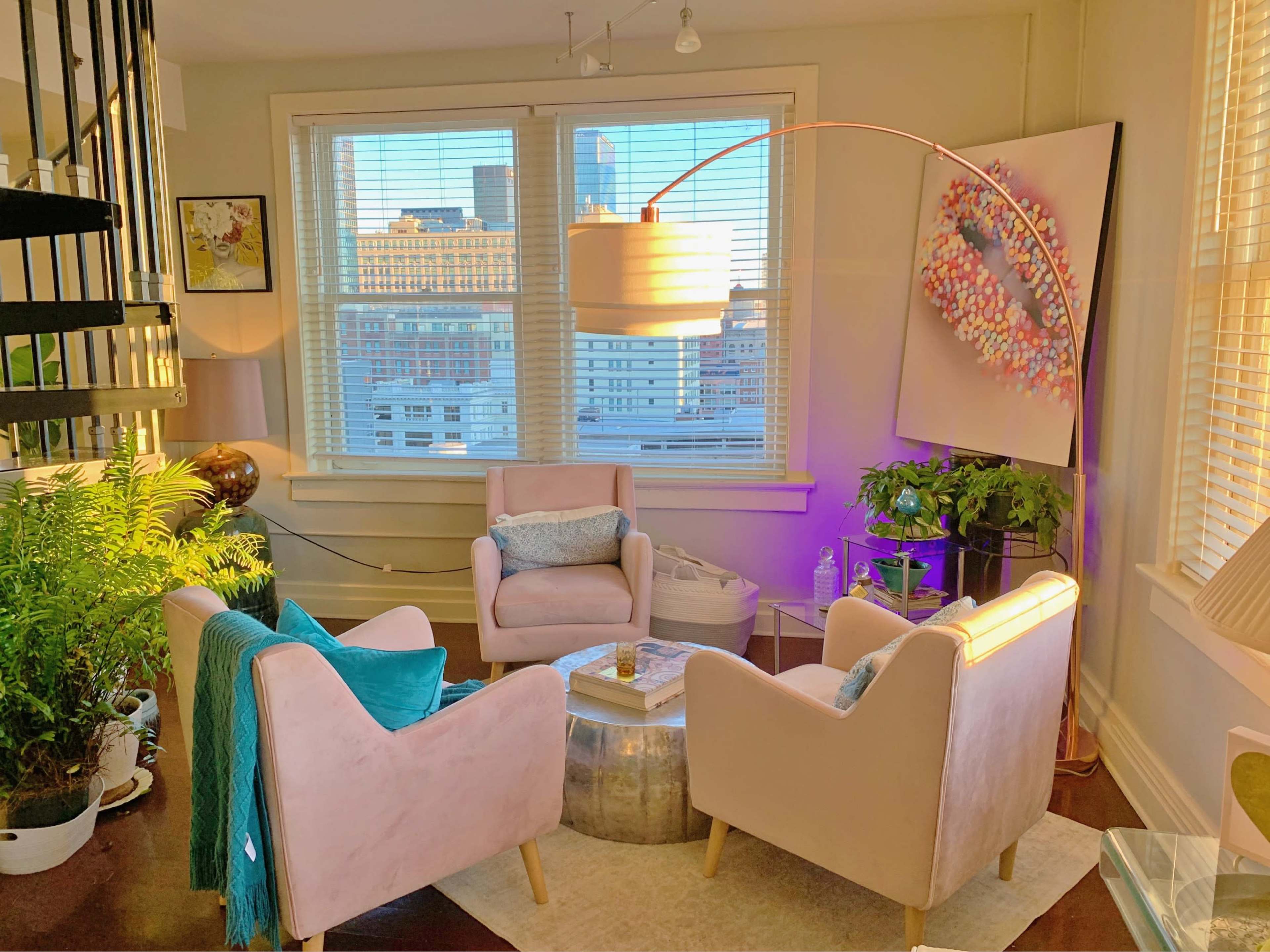 The scene features a cozy living area with two pink chairs, a round table, and a large window showcasing a city view, illuminated by a modern floor lamp.